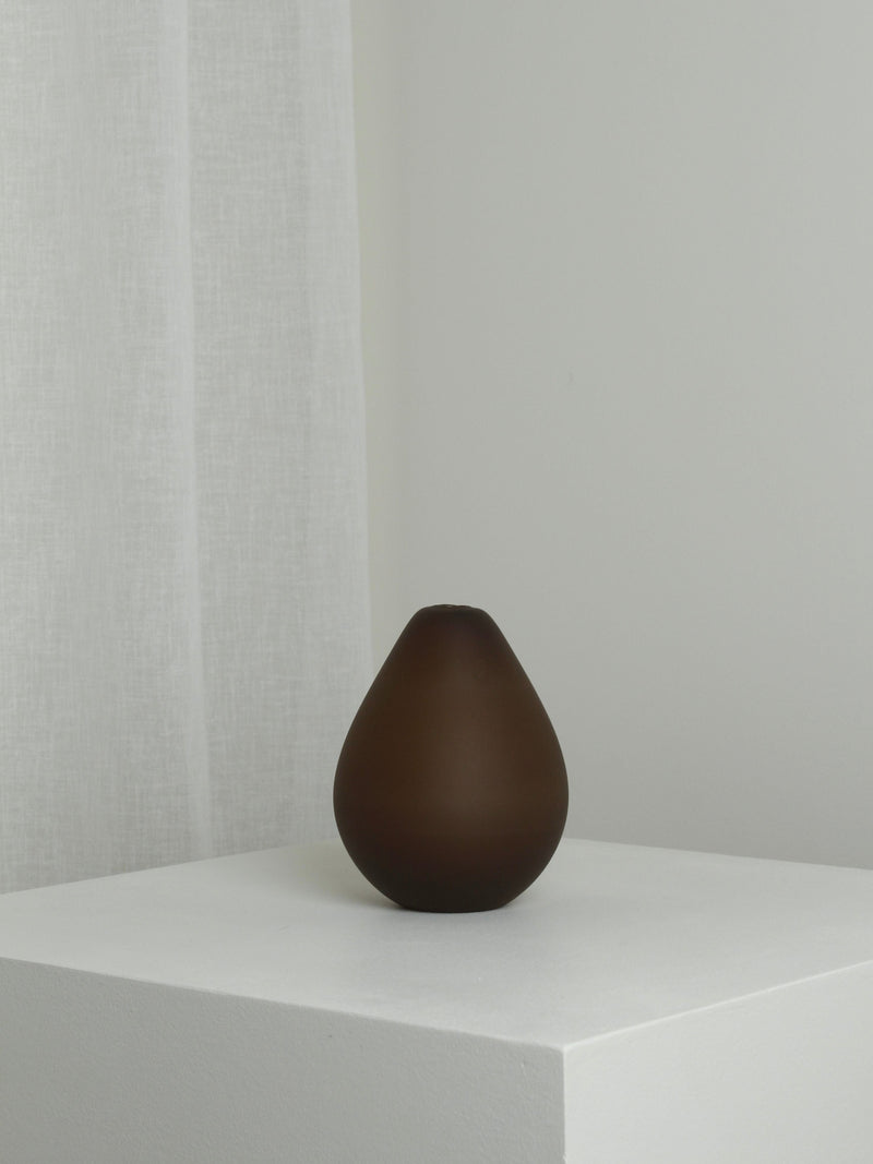 The Medium Vase in Chocolate by Verre d'Onge, round and elegant, is displayed on a white pedestal in a minimalist setting with a sheer white curtain behind it.