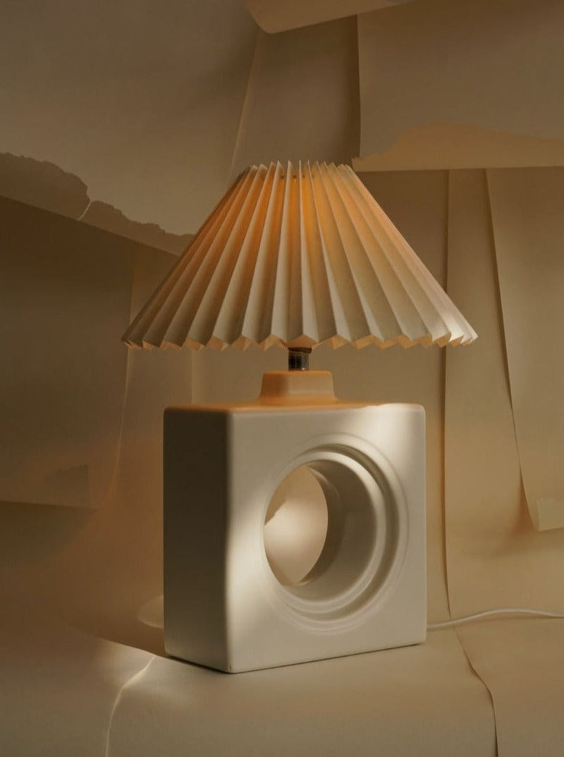 The Modernist Table Lamp by Les Objets Amsterdam features a pleated paper shade and a square ceramic base with a large circular hole, shown on a soft beige background with layered paper sheets.