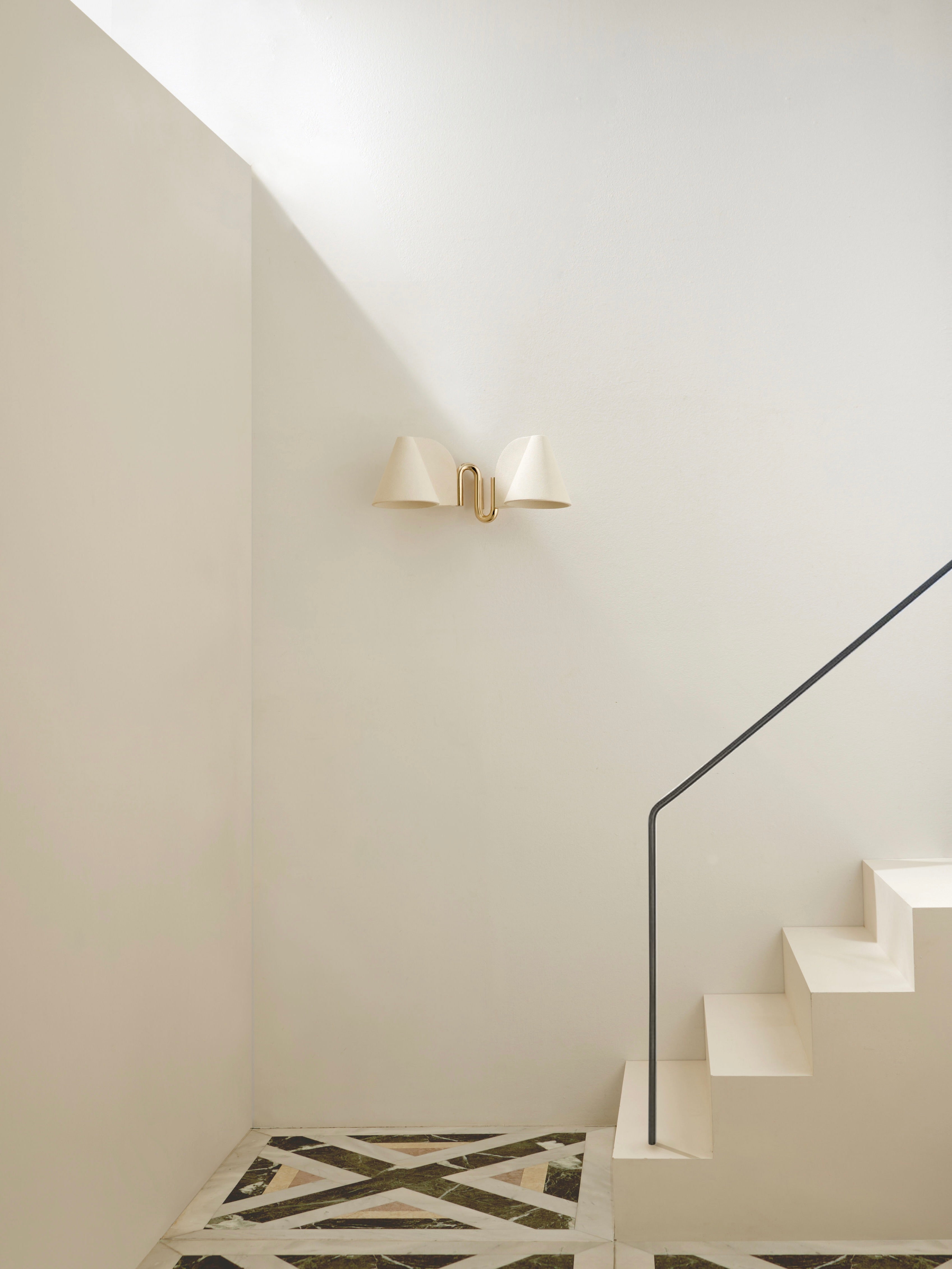 A minimalist staircase with white steps and a black handrail stands against a light wall. Above, the 'Synapse' Wall Light by Mariza Galani adds a contemporary brass accent, while the floor displays a geometric marble pattern.