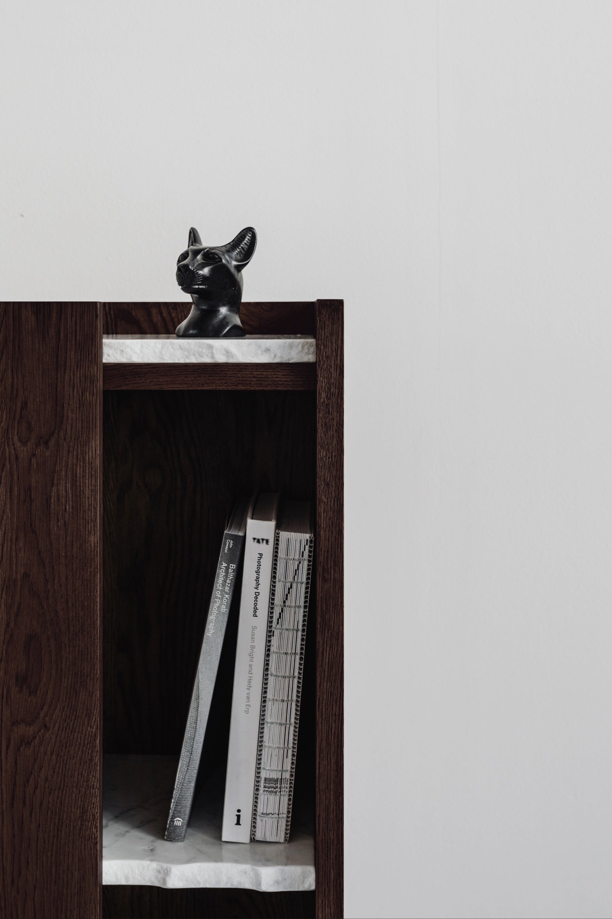 The PAVO Cabinet by un’common, a dark solid wood console with a white marble shelf, holds books and is topped with a black cat figurine, standing against a plain white wall.