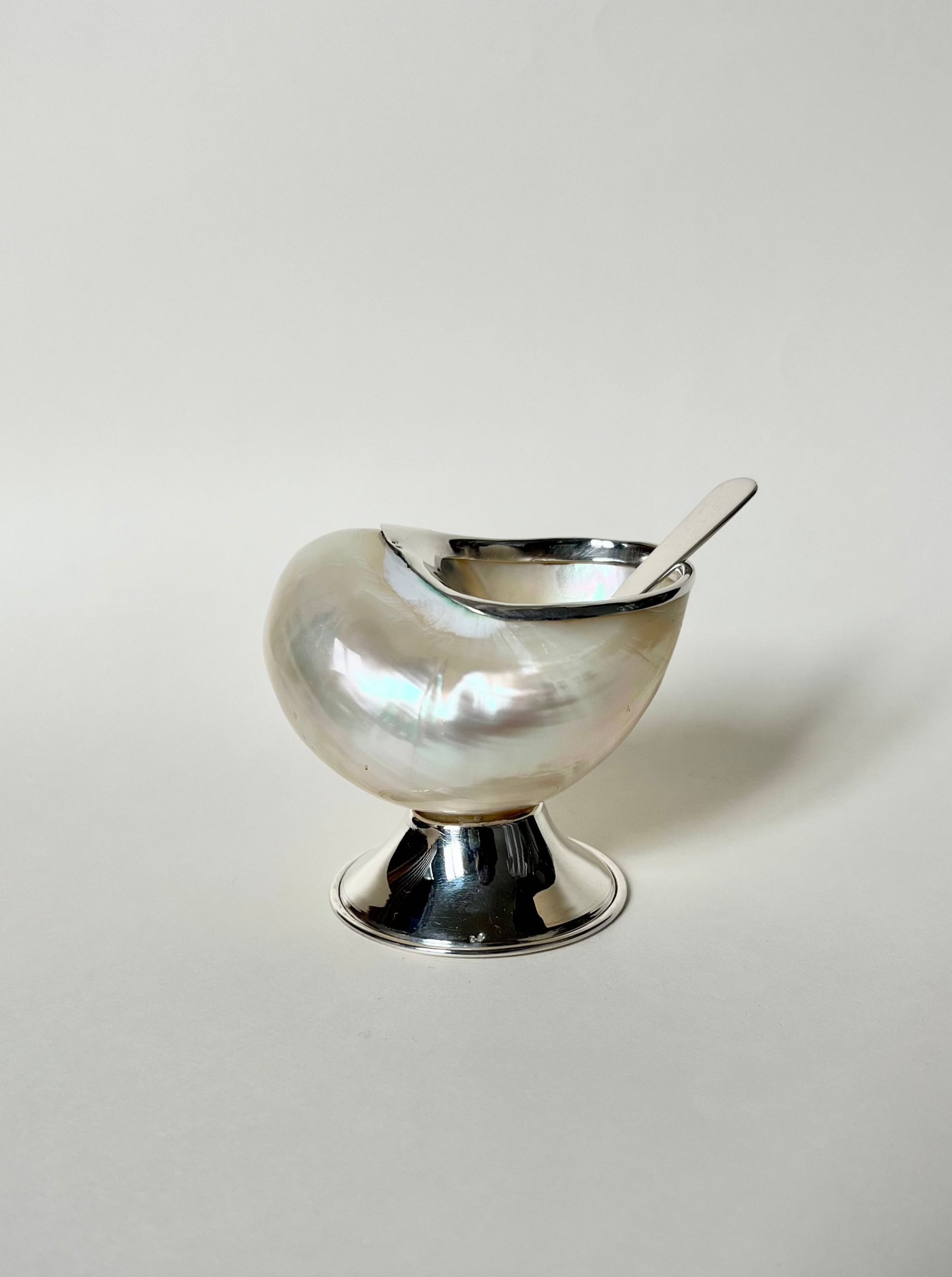 A polished silver and mother-of-pearl Shell Sugar Bowl by Tavares 1922, with matching spoon, sits on a reflective pedestal—adding charm to an elegant tabletop against a plain light background.