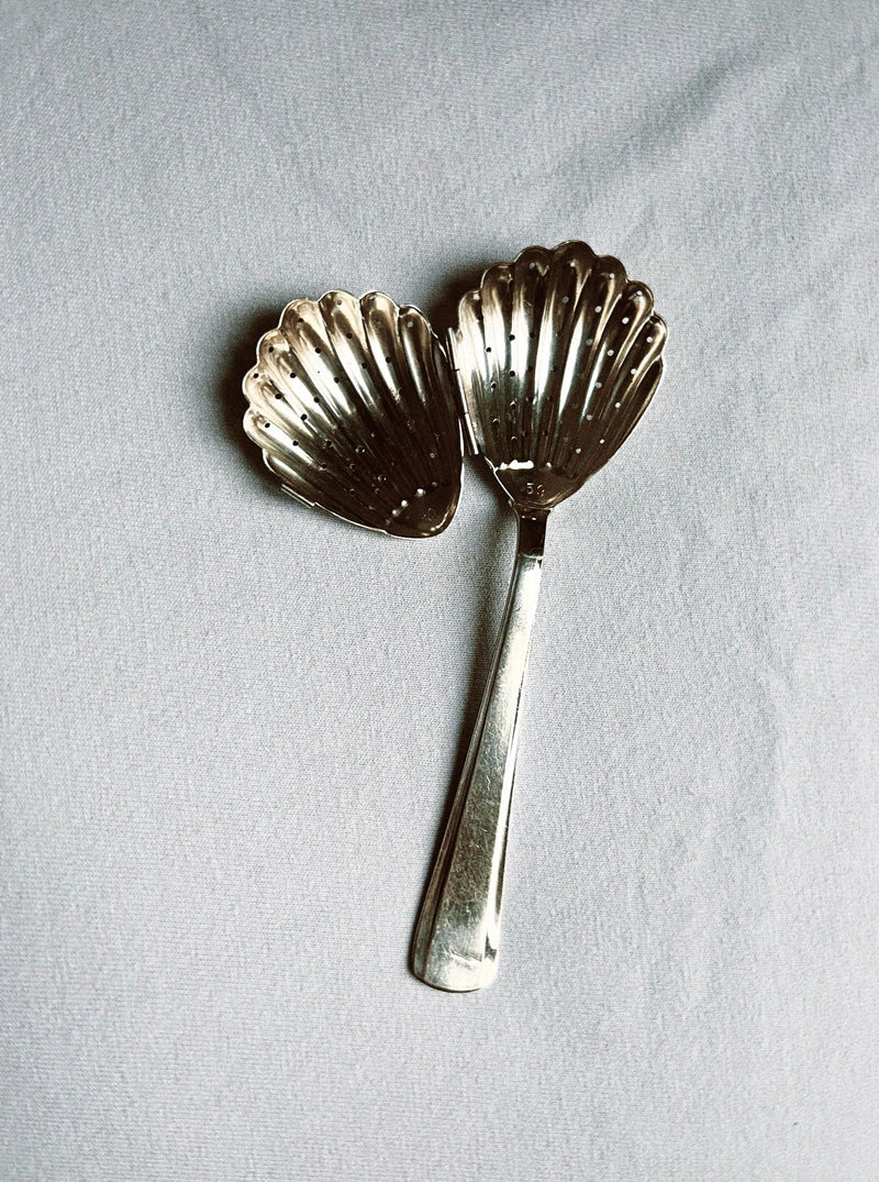 The Manushi Shell Shaped Tea Leaf Infuser, featuring two scallop shell-shaped heads in silver, is displayed on a light gray fabric background.