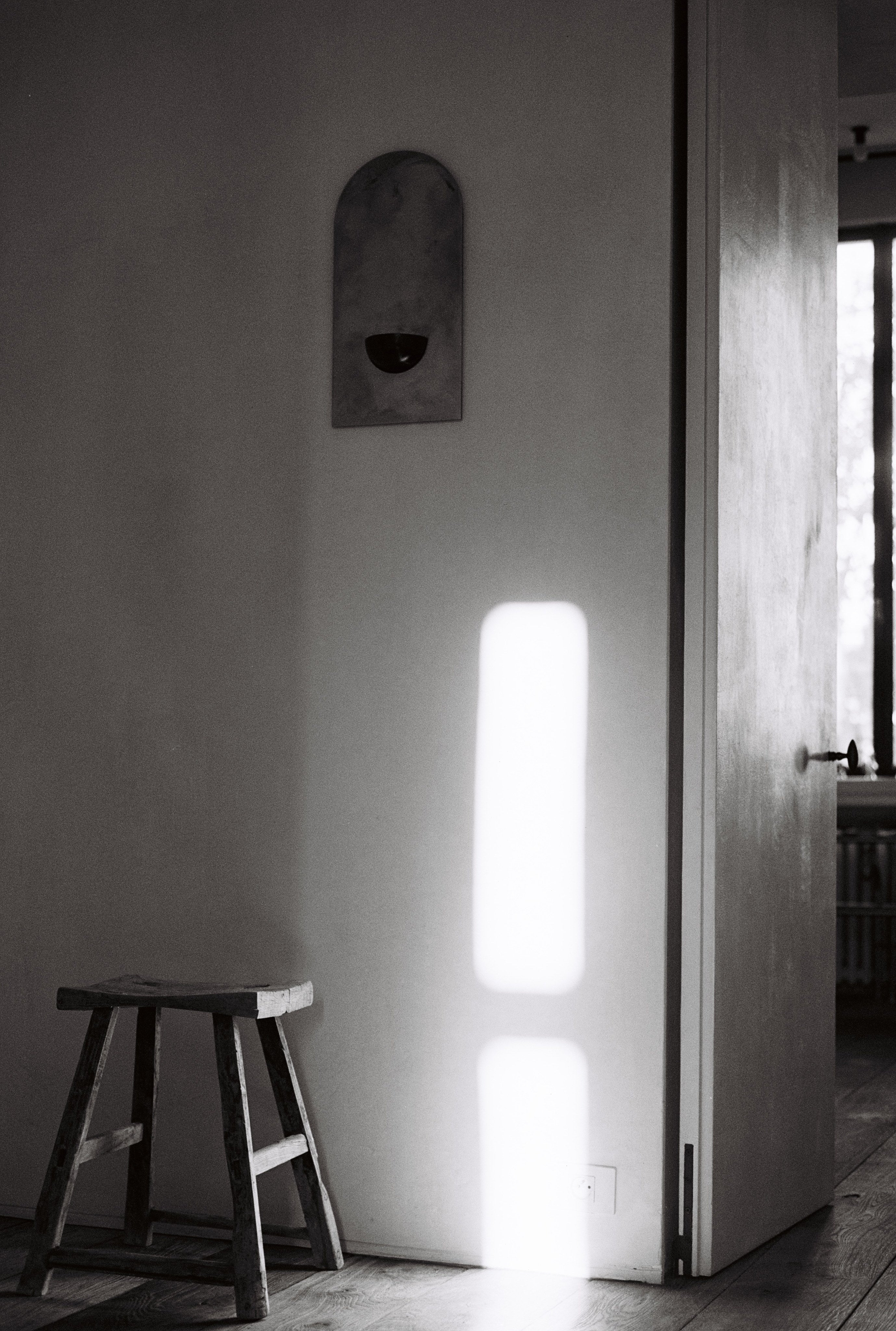 A shaft of sunlight illuminates a white wall and wooden floor in a minimal room. The COSEINCORSO "Mirror of Simple Souls"—a contemporary steel mirror—pairs with a rustic stool, adding character to the minimalist decor.