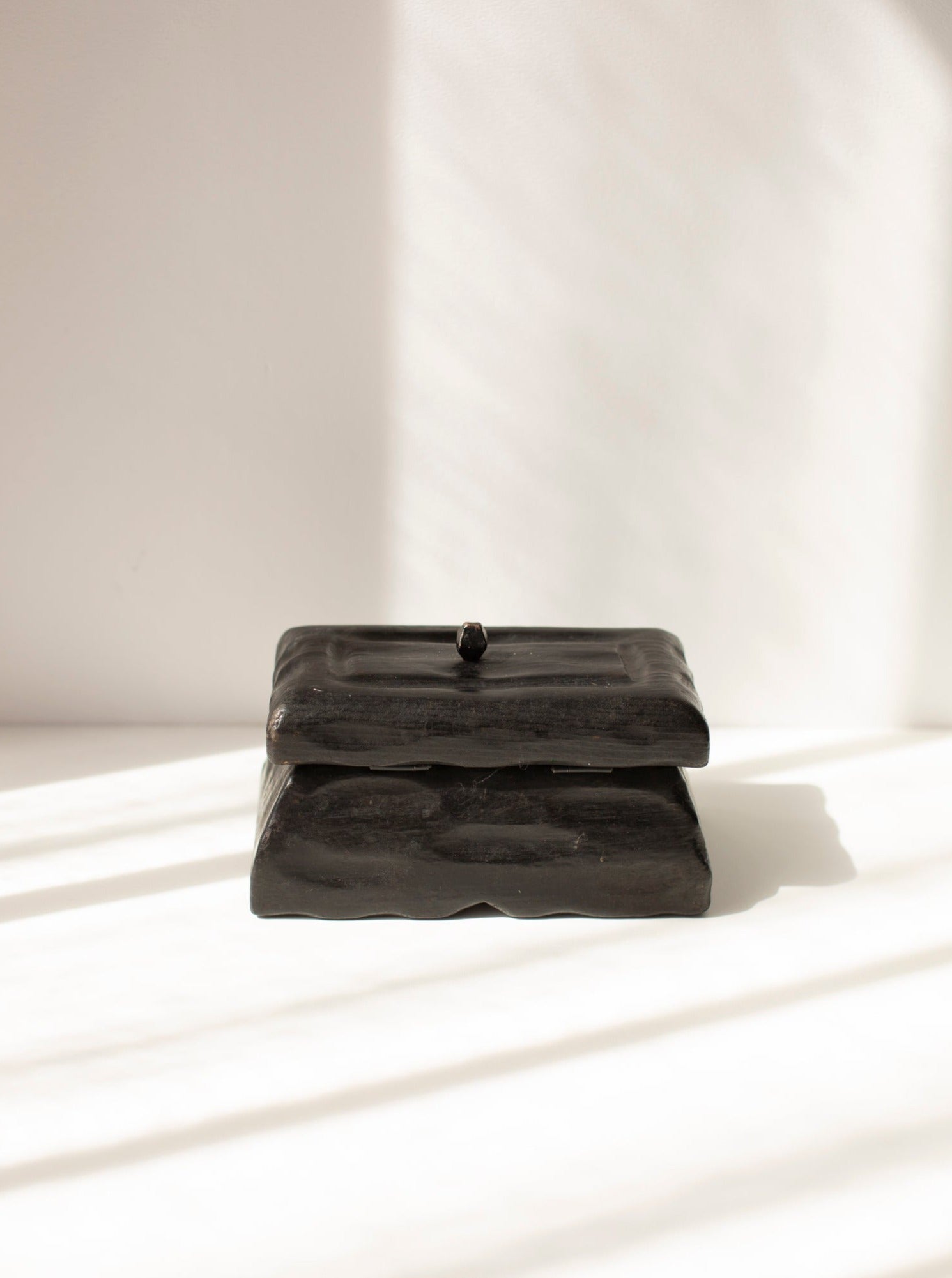 A square, black wooden handmade box with a lid sits on a white surface. The **Black Wooden Handmade Box** by **CU-RA-TED** has a smooth, glossy finish with minimalist design. Sunlight casts soft, diagonal shadows across the scene, adding depth and contrast—perfect for storing small items.