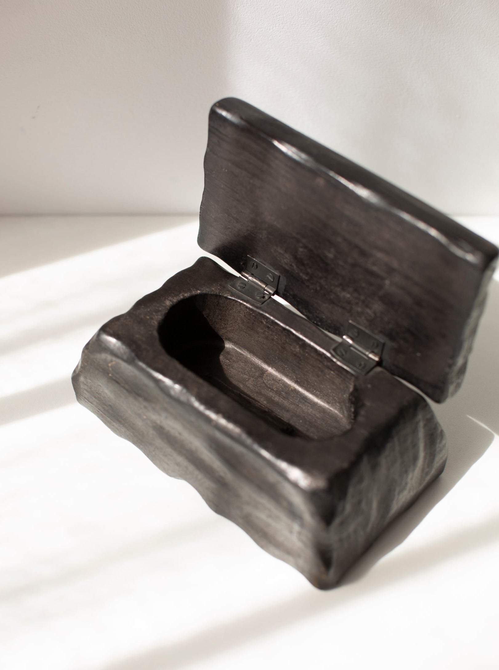 A small, Black Wooden Handmade Box by CU-RA-TED made of a rough material is open on a white surface. The interior of the box is empty and its lid is attached with two metal hinges. The sunlight casts soft shadows on the surface, highlighting the premium quality wood used in its construction.