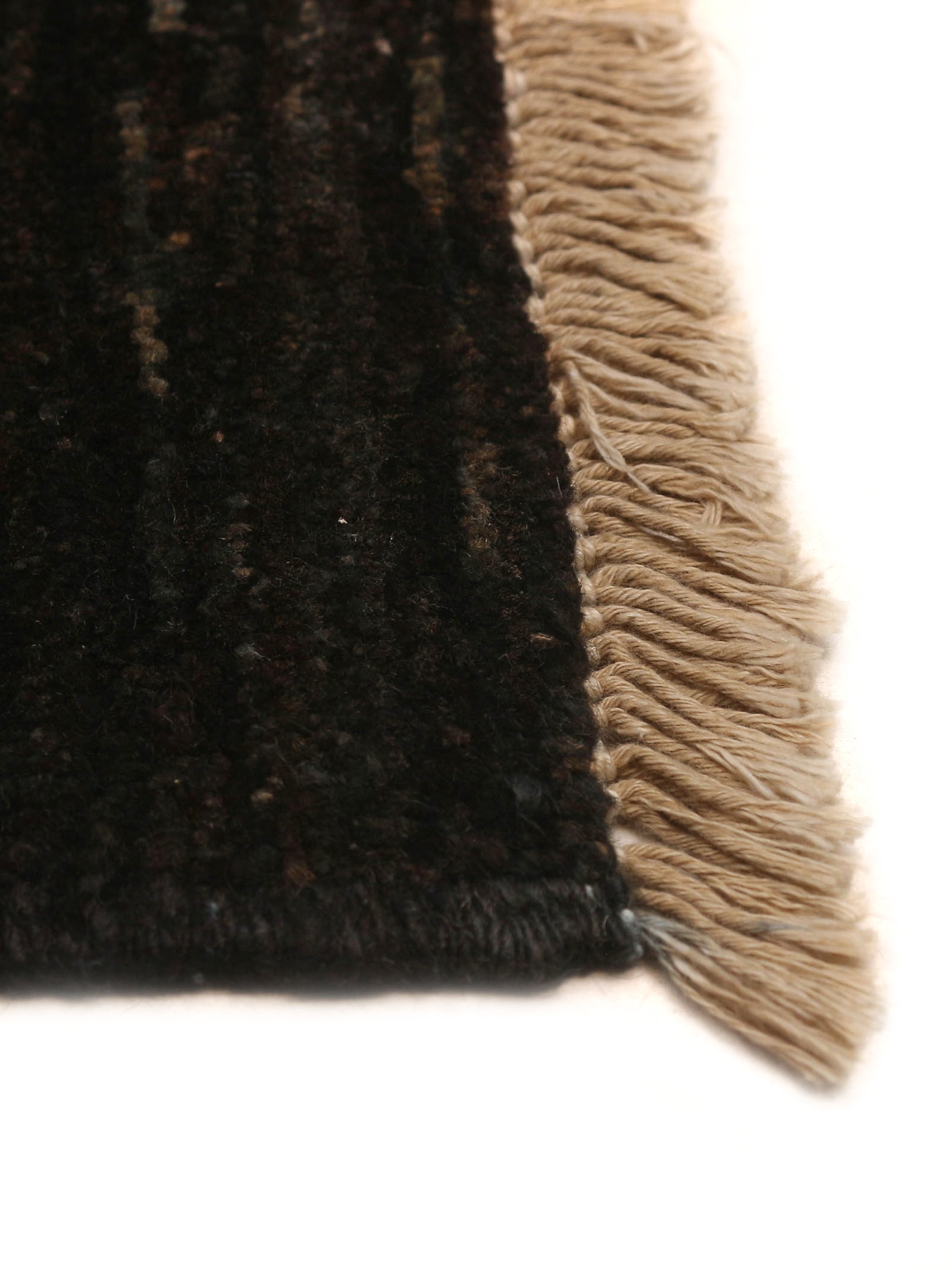 Close-up of a corner of the Ziegler Rug in Obsidian (247 x 277 cm) by catalog:three, showcasing its dark, textured wool with light-colored fringe. The hand-knotted rug's woven details contrast beautifully with the smooth fringe.