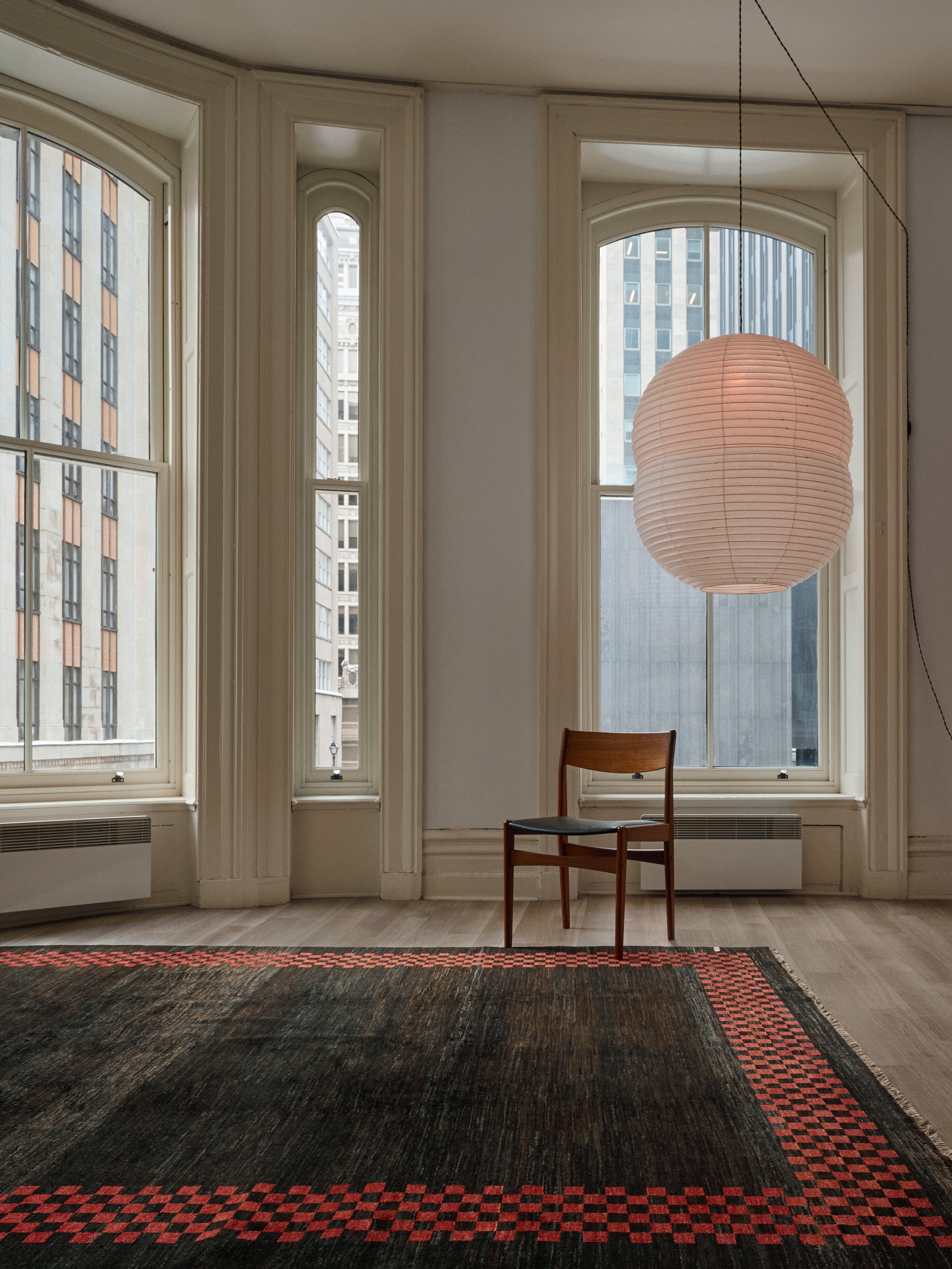 A minimalist room with large arched windows features a single wooden chair and a paper lantern overhead. The floor showcases the Ziegler Rug in Obsidian by catalog:three, with a dark tone and red checkered border, while urban buildings are visible beyond the glass windows.