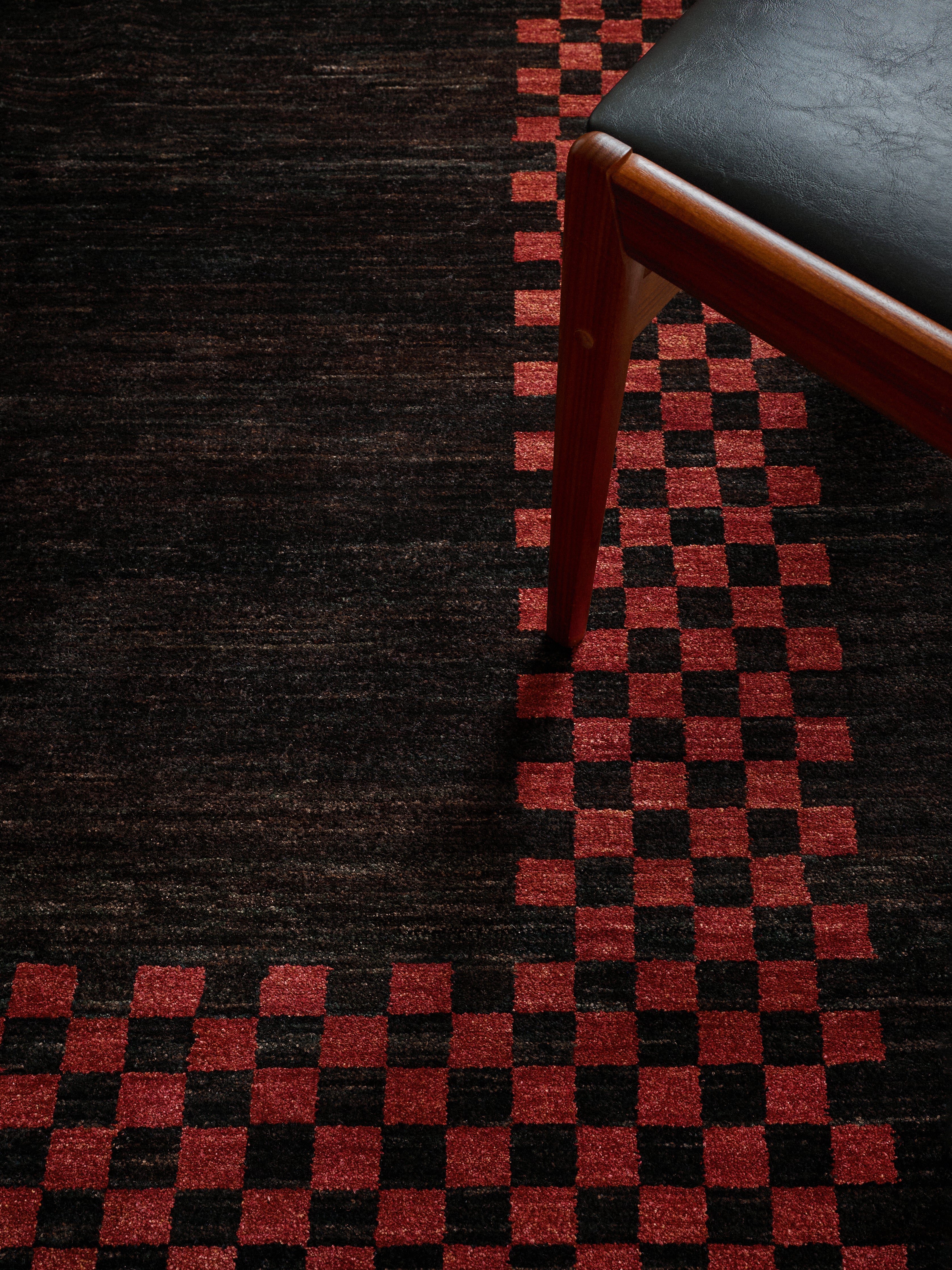 A Ziegler Rug in Obsidian from catalog:three, featuring a red checkered pattern, has a wooden chair with black upholstery positioned in the upper right corner. The moody lighting enhances its unique charm.