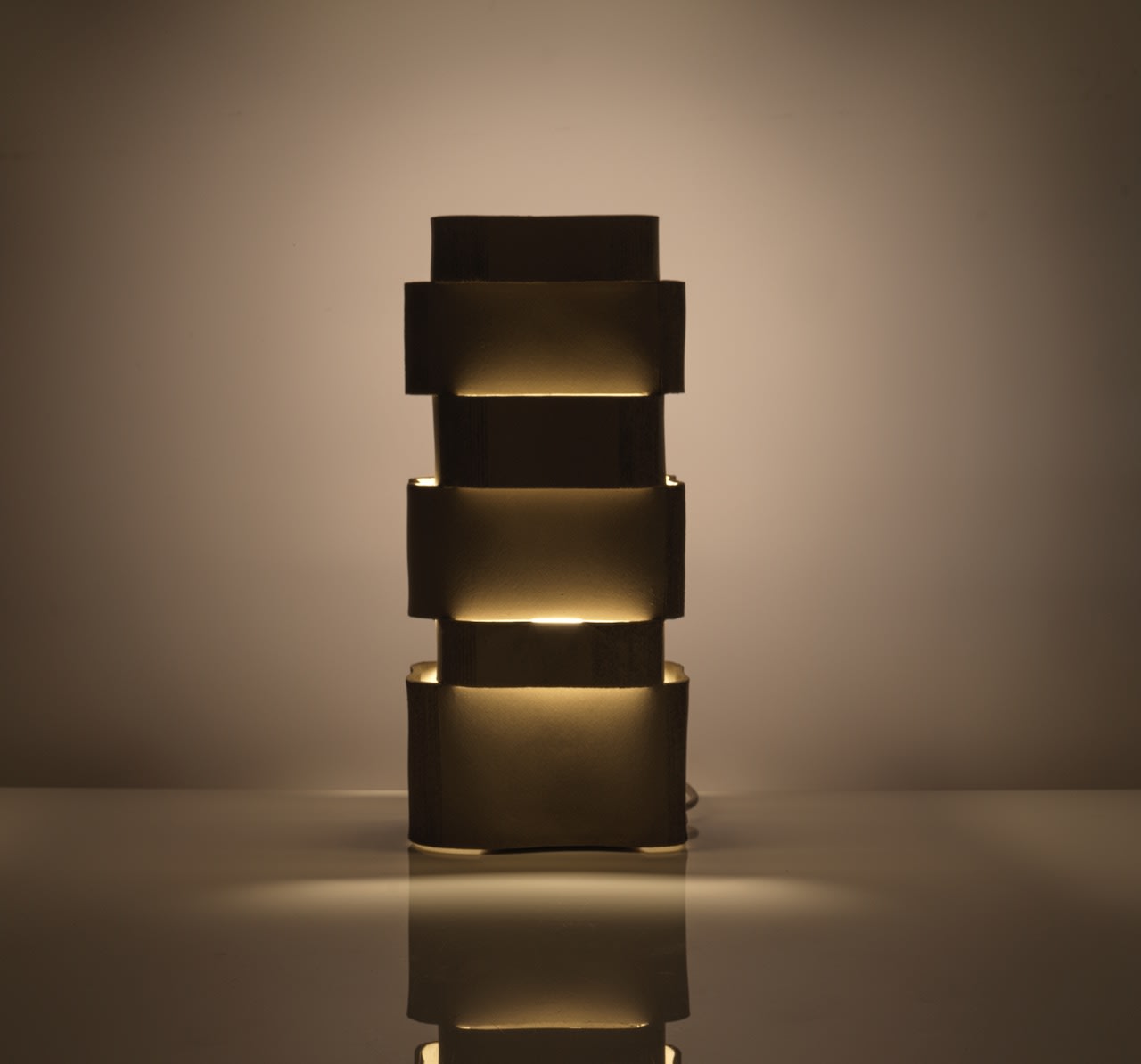The Katia Langeard Too 60-2 Table Lamps in Ceramic feature a modern rectangular design with three stacked, offset segments, emitting warm, soft bedside lighting that casts gentle shadows and reflects on glossy surfaces.