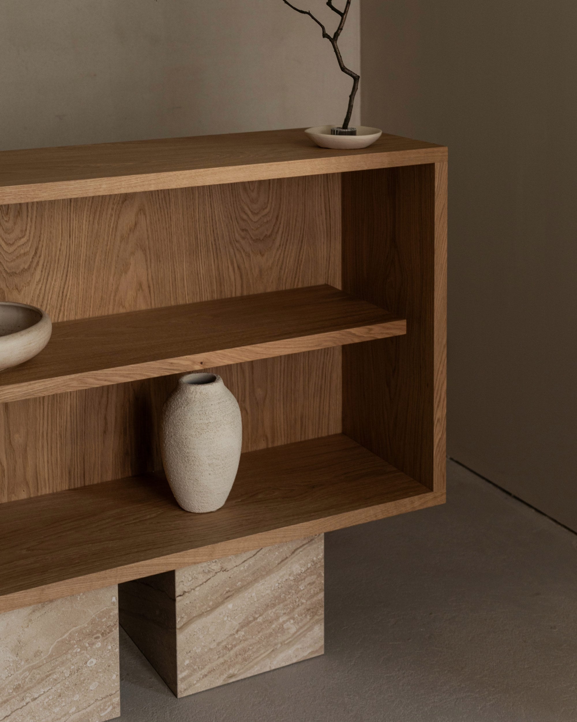 The BLOCK Sideboard by un'common, featuring minimalist wood and a light stone base, elegantly displays a white bowl, ceramic vase, and bare branch dish against a plain beige wall and floor.