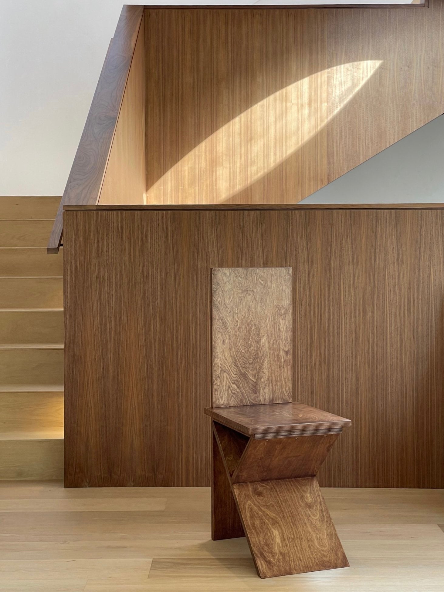 The Flat Pack Chair by Goons from GOONS, featuring minimalist geometric angles, is positioned before a staircase and a wood-paneled wall, bathed in natural light.