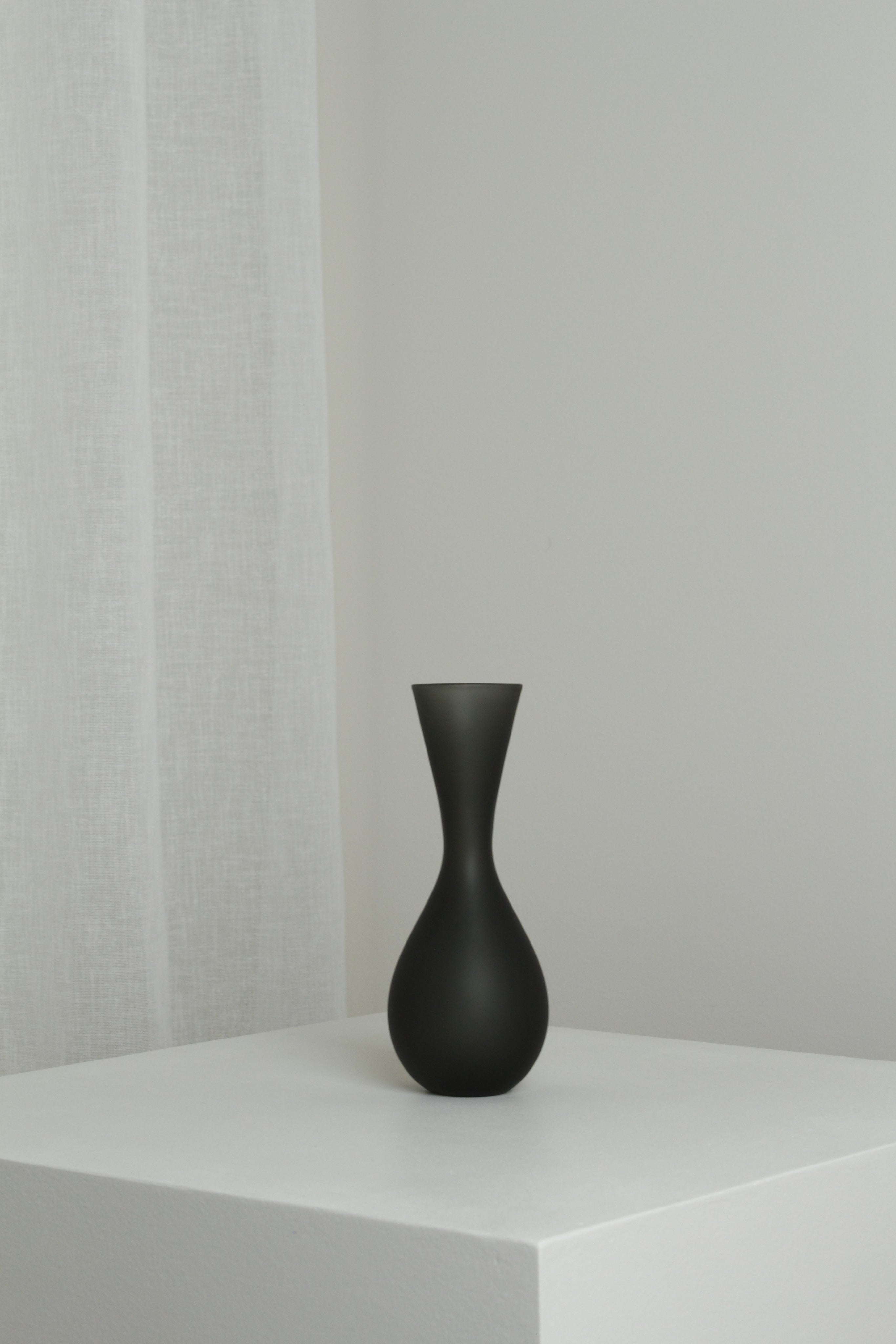 The Verre d'Onge Medium Vase in Smoke, with an hourglass shape and matte black finish, sits on a white pedestal beside a white curtain against a light gray wall in a minimalist setting.