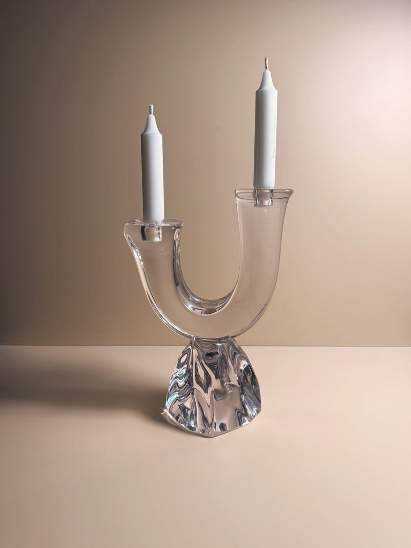 A Solid Crystal Glass Brutalist Candelabra by Daum (1950s) from Dodo Vintage, featuring two white taper candles, sits on a smooth beige surface against a neutral background.