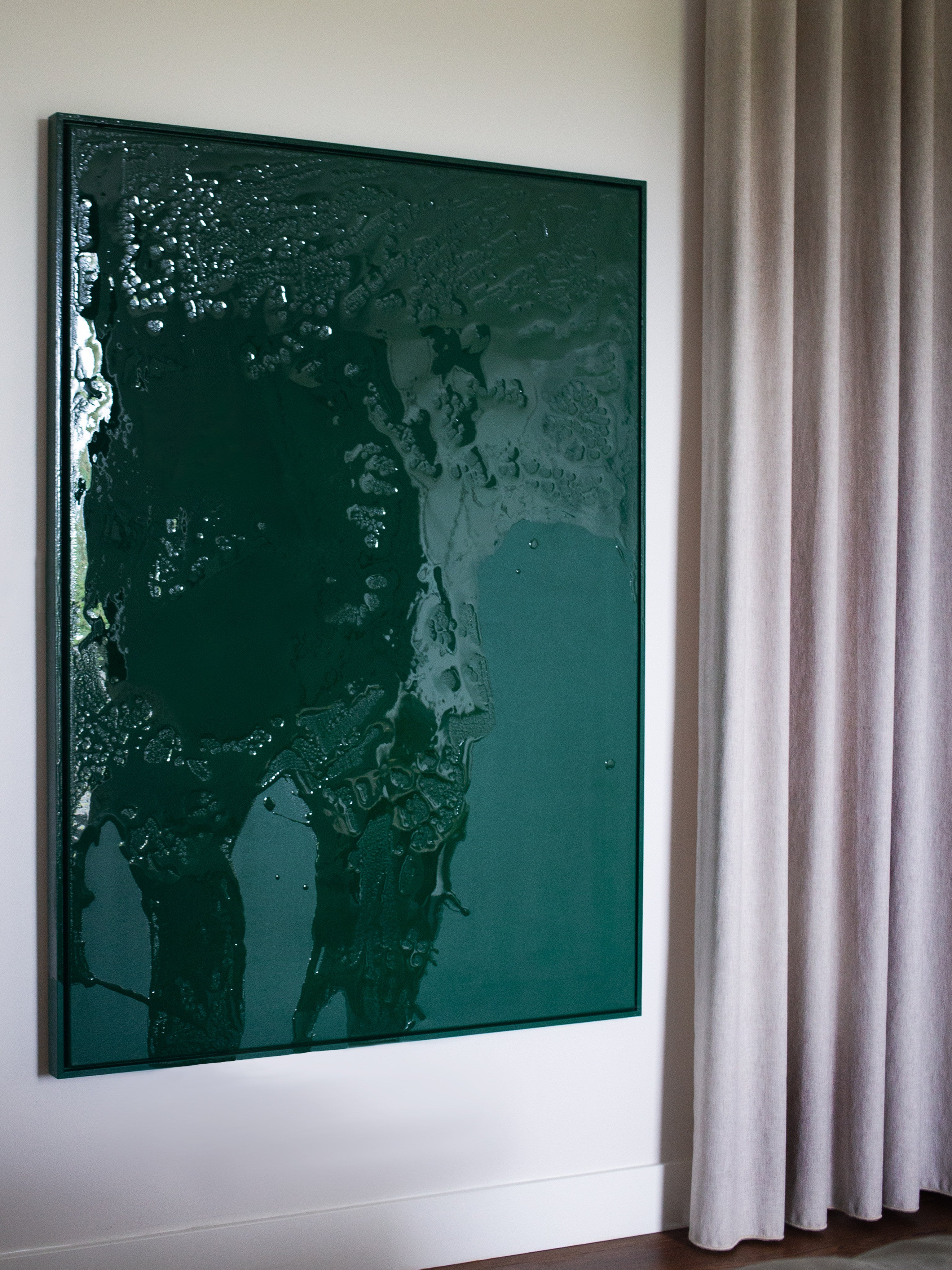 Green Flux - Uncategorized" by STUDIOPOLS, a large glossy dark green acrylic and epoxy resin wall art, hangs on a white wall beside beige curtains. Its textured surface catches and reflects light, adding modern elegance to the room.