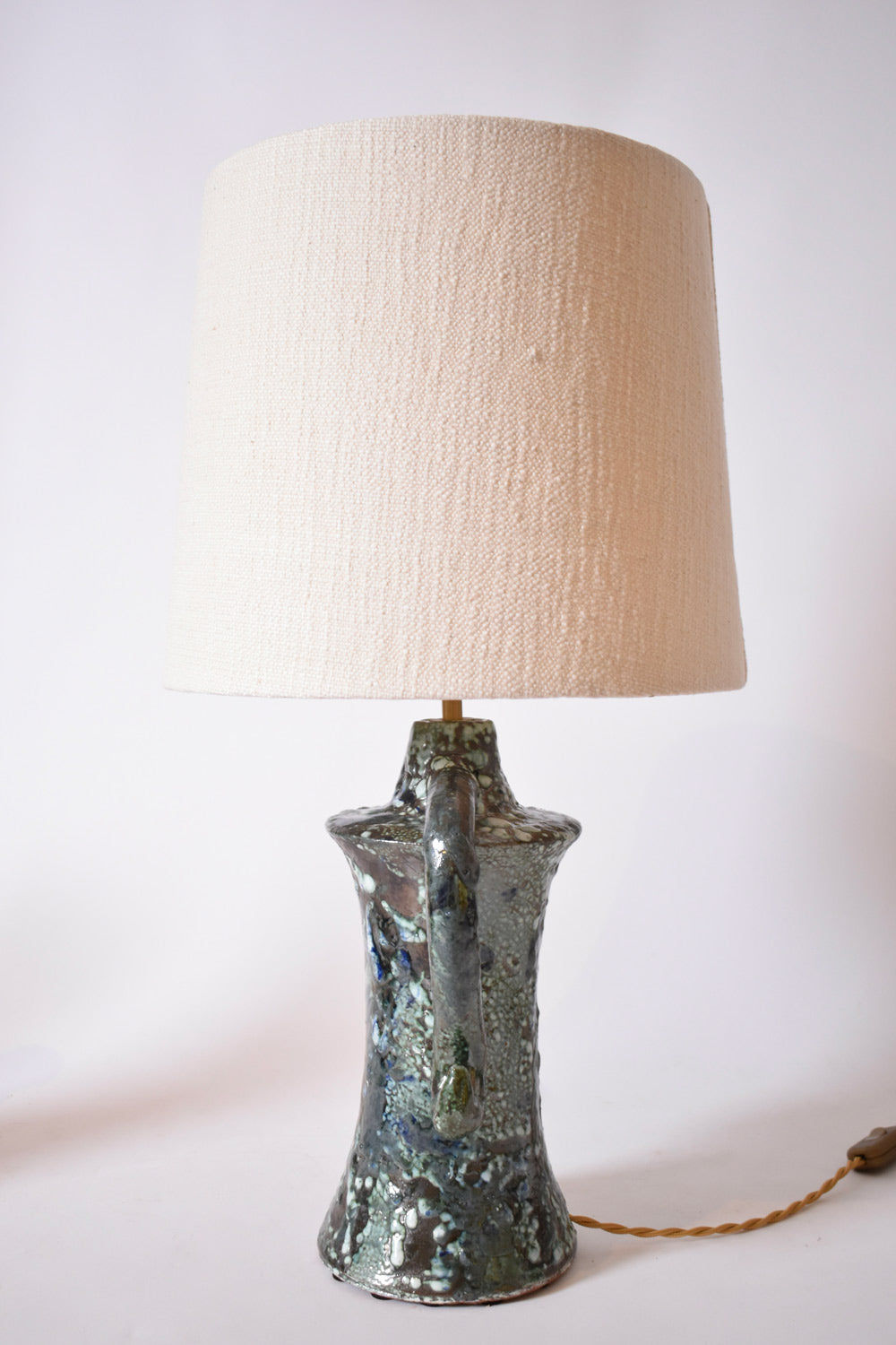 The "Kasaï" Erato Blue and Green Ceramic Table Lamp by Barracuda Interiors features a textured, multicolored base and a large off-white fabric shade. Its visible cord adds a relaxed touch, ideal for contemporary spaces with plain white walls.