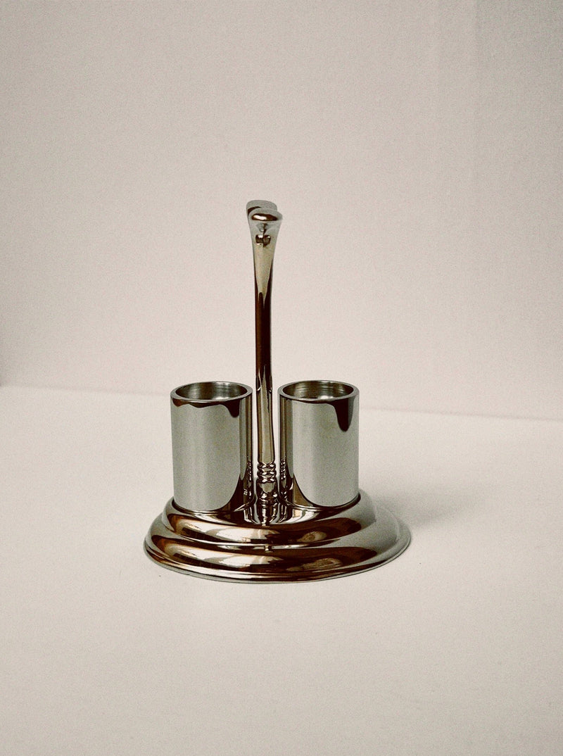 The Manushi Salt & Pepper Shakers in Holder feature a shiny metal design with two cylindrical compartments and a central handle, displayed on a plain white surface against a light background.