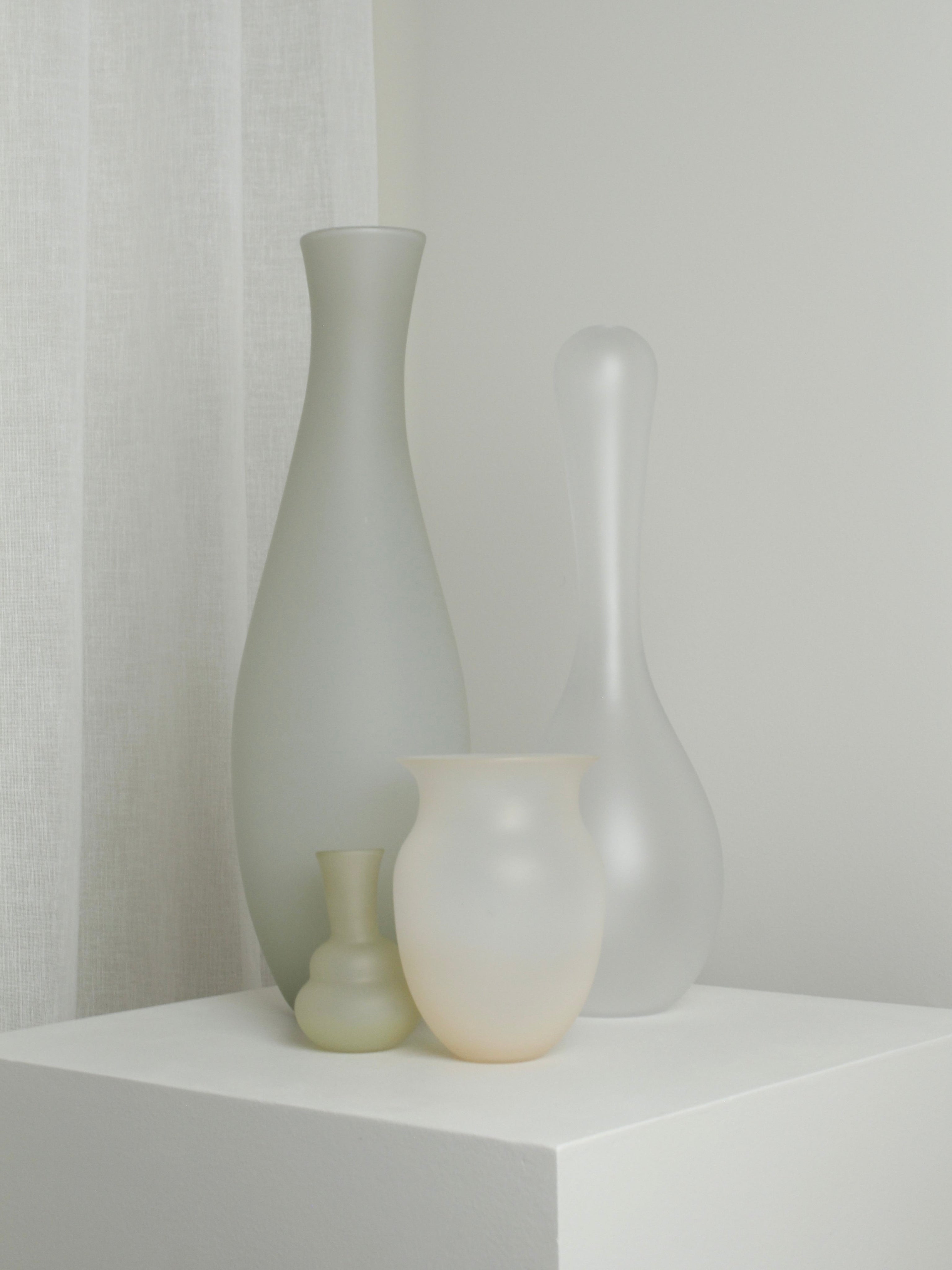 Four frosted glass vases, including Verre d'Onge's XLarge Vase in Ghost, are displayed on a white pedestal in a softly lit, minimal room with white walls and a sheer curtain.