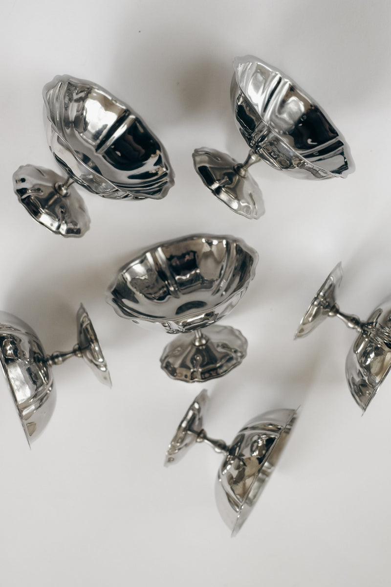A set of six vintage stainless steel ice coupes by septembre studios is arranged on a white surface—some upright, others tilted or upside down—creating an intriguing pattern with their reflective finish.