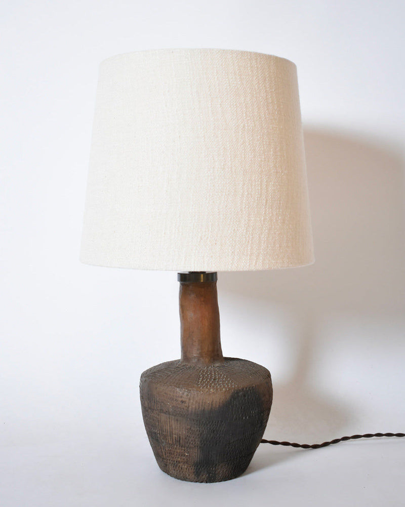 The African Brown Ceramic Table Lamp by Barracuda Interiors (1970s) features a textured dark brown terracotta base with a subtle geometric pattern, a large off-white fabric shade, and a twisted brown cord visible on a white surface.