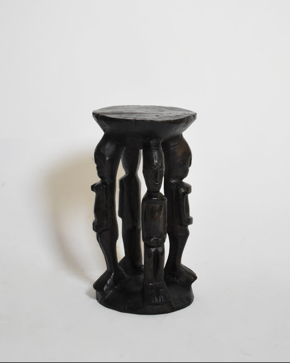 The African Anthropomorphic Side Table (1960s) by Barracuda Interiors is crafted from carved ebonised wood, with four upright figures as legs supporting a round seat, shown on a plain white background.