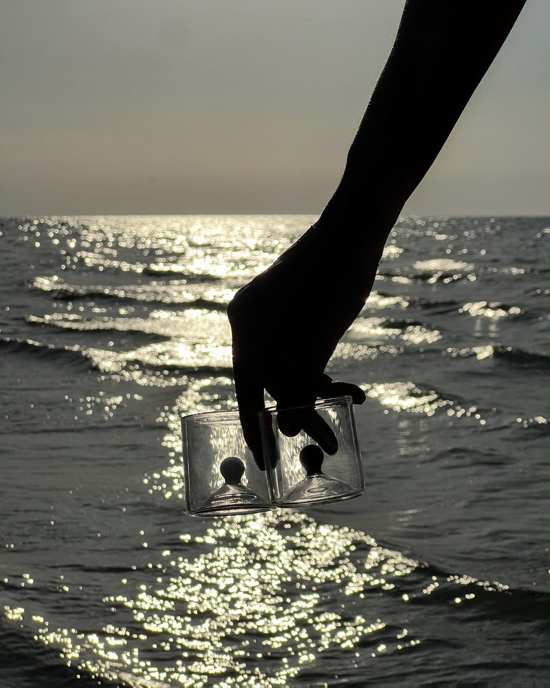 Hand holding green glass shot duo set against sparkling ocean water