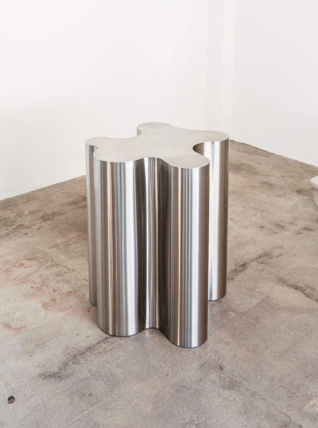 The Silver Root - Tall side table by Caia Leifsdotter, with its sculptural steel form and polished silver finish, adds a modern touch to a minimal white-walled room with a bare concrete floor.