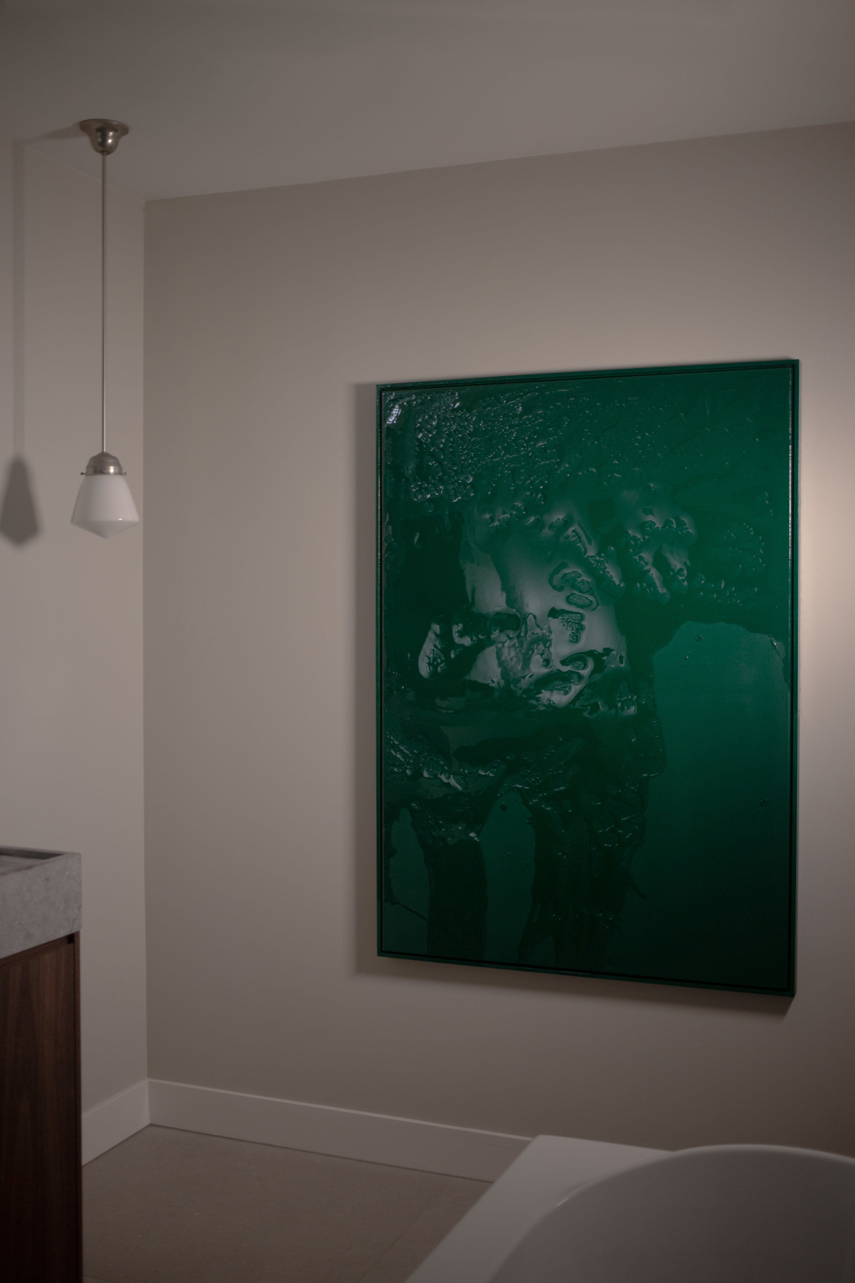 A minimalist bathroom features Green Flux by STUDIOPOLS, a striking green acrylic and epoxy resin artwork on the wall, with soft overhead lighting, white walls, and a glimpse of a wooden vanity and bathtub in the foreground.