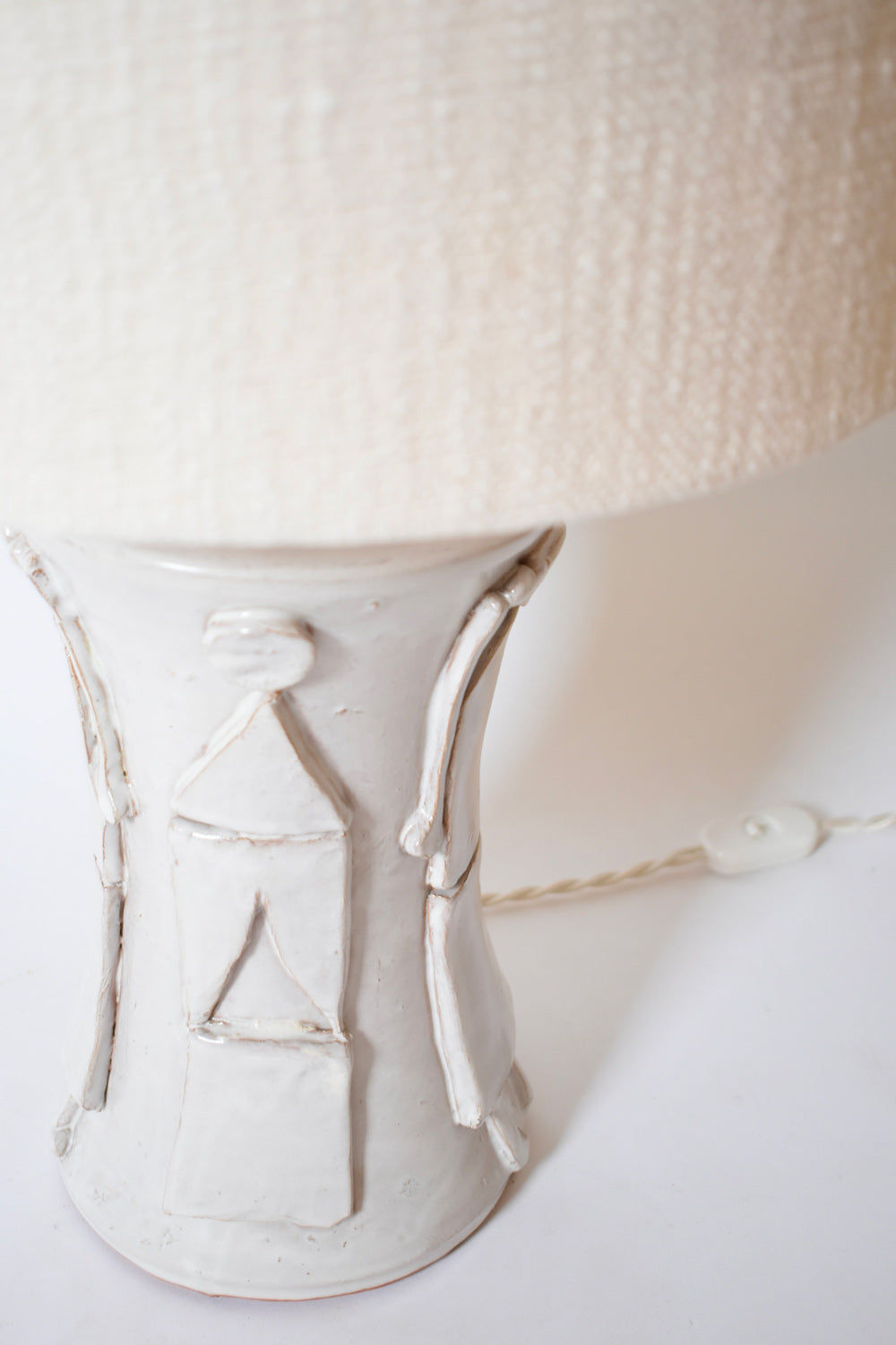 A close-up of the "Kasaï" Avignon Ceramic Table Lamp by Barracuda Interiors shows its off-white base with abstract raised patterns and a textured shade—ideal for customizable decor. A twisted cord with inline switch appears in the background.