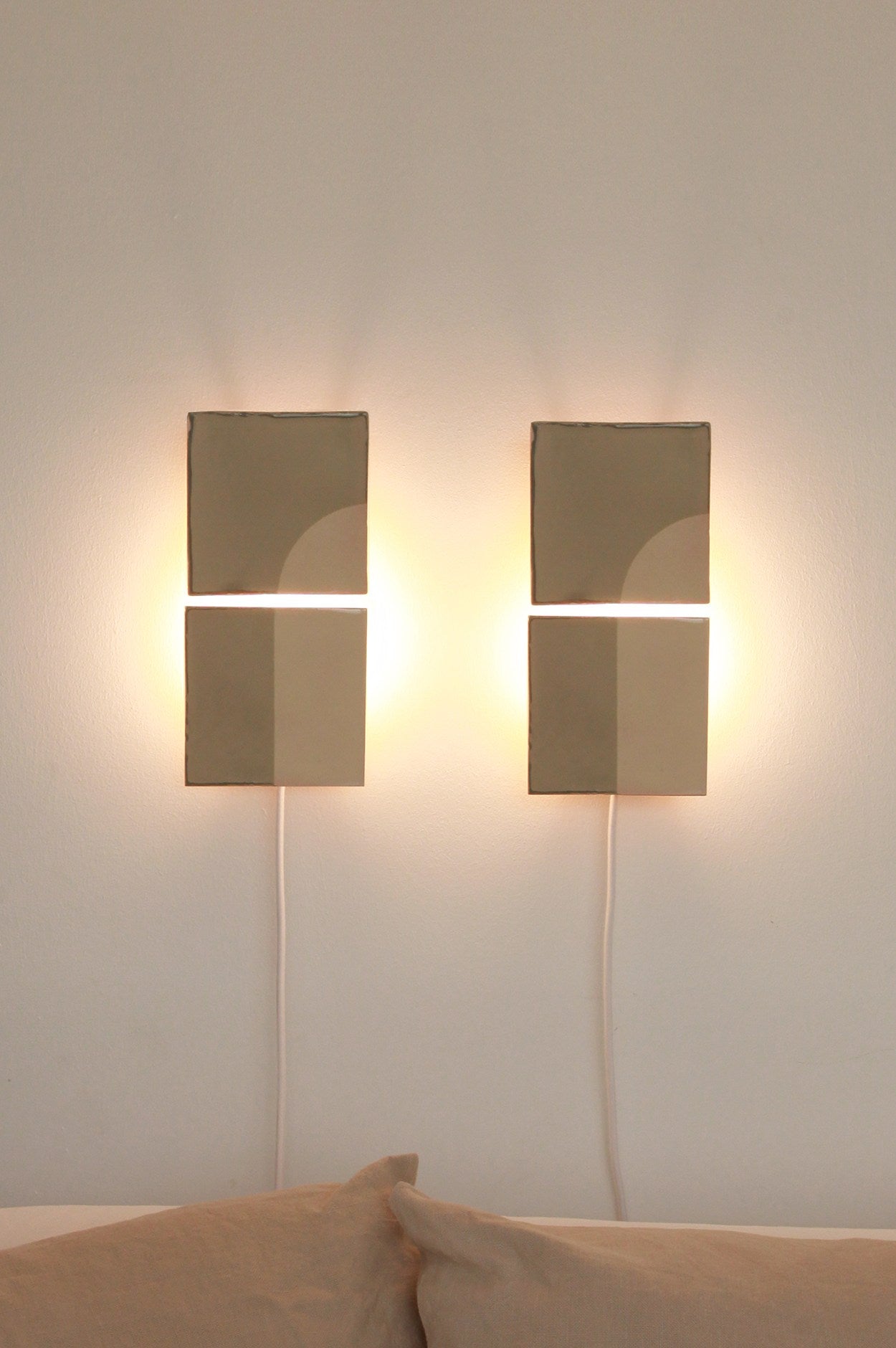 Two Tiles Door V ceramic wall appliques by Violaine d'Harcourt, with geometric patterns, emit warm light above a beige sofa. White cords hang against a light wall, highlighting their artisanal craftsmanship.