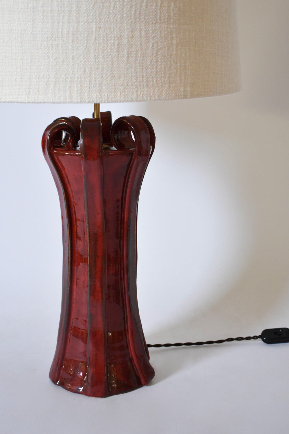 The "Kasaï" Henry Red Ceramic Lamp by Barracuda Interiors features a glossy, sculpted base, large off-white fabric shade, and twisted black cord with inline switch, shown against a plain white background.