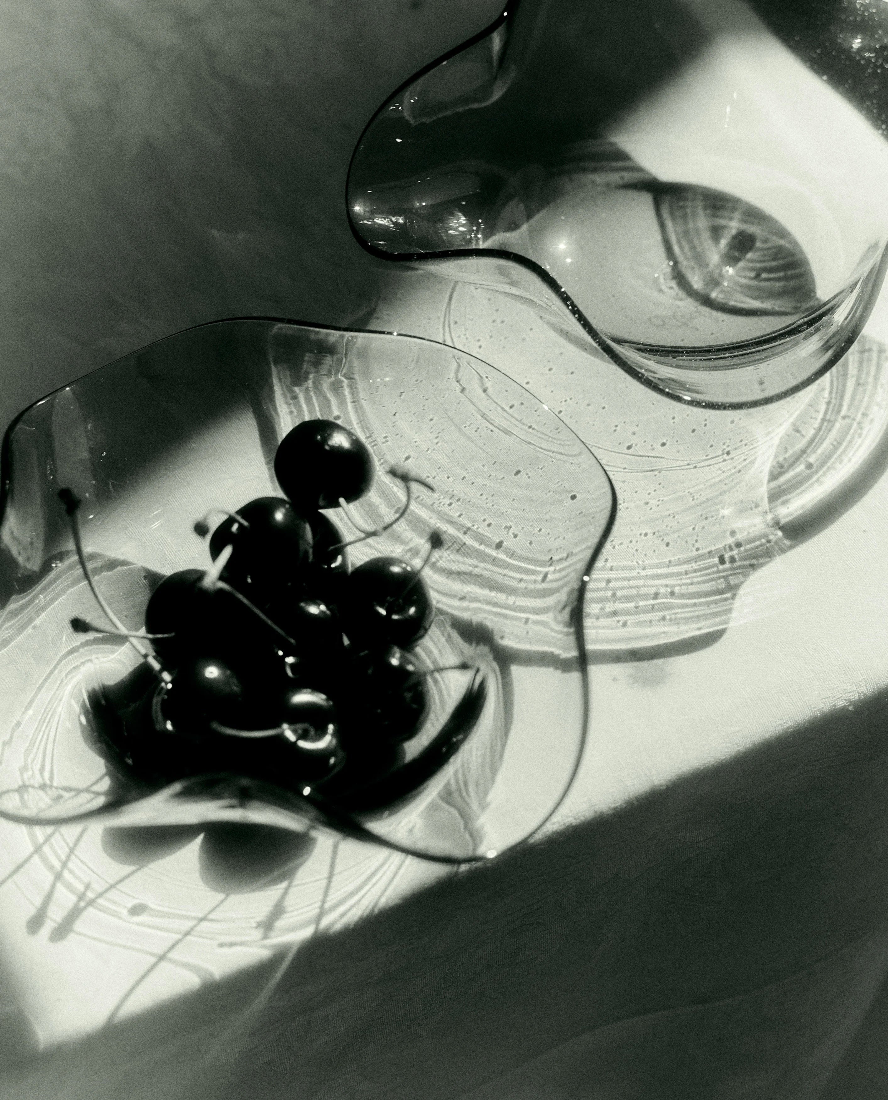 A black-and-white photo of the Michele Bowl Small by Akua Objects, its curved edges filled with cherries, sits on a sunlit surface casting dramatic shadows and reflections.