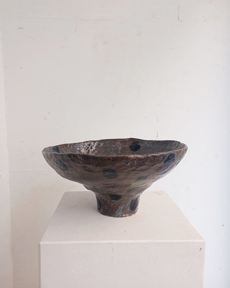 The Coupe 40 by Claire Pain is a handmade dark ceramic bowl with an uneven rim and black spots, displayed on a light pedestal against a plain white wall.