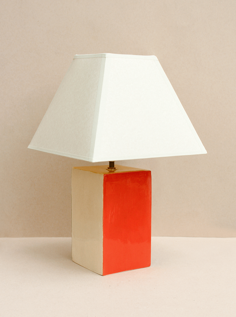 The Paloma Lamp by Casa Veronica is a hand-built ceramic lamp featuring a custom square shade and a rectangular base, divided into cream and bright red, set against a beige background.