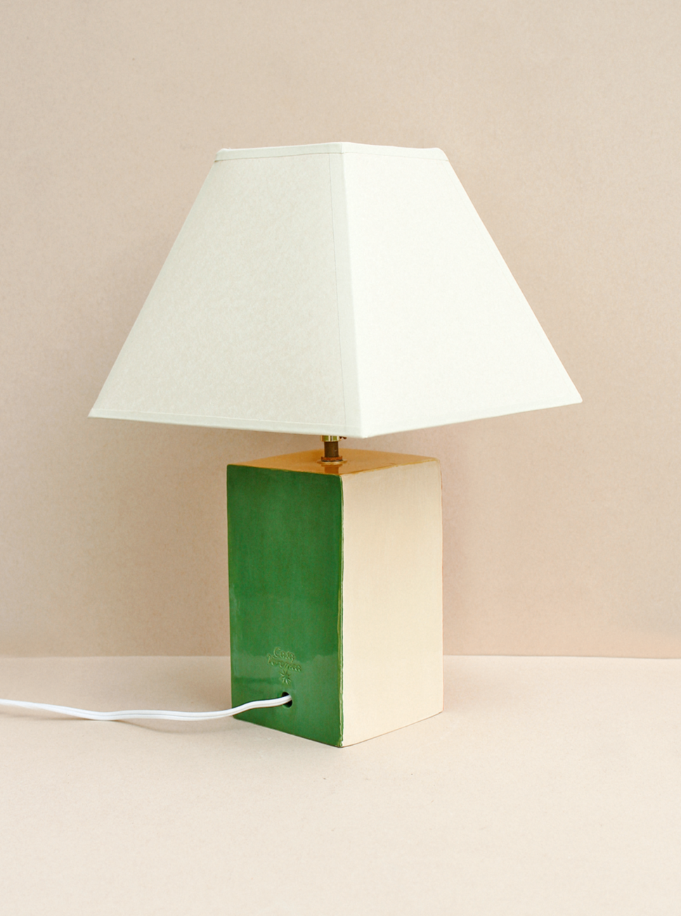 The Paloma Lamp by Casa Veronica features a hand-built ceramic rectangular base painted half green and half beige, a custom square lampshade, and a white power cord plugged into its base, resting on a light surface.