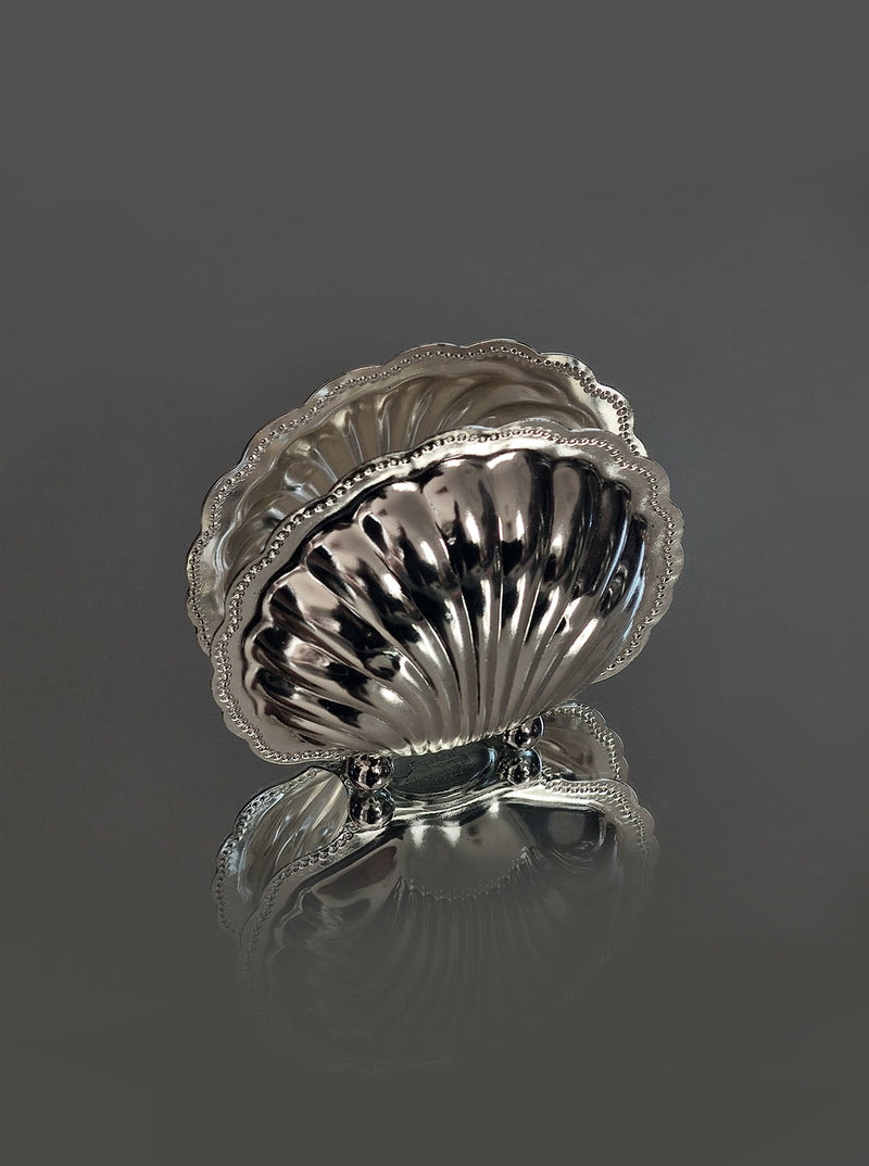 The Les Objoies Napkin Shell Holder features a polished silver-plated finish with ornate scallop shell details and decorative feet, making it ideal for seaside-inspired tabletop decor. Shown on a smooth gray surface against a plain background.