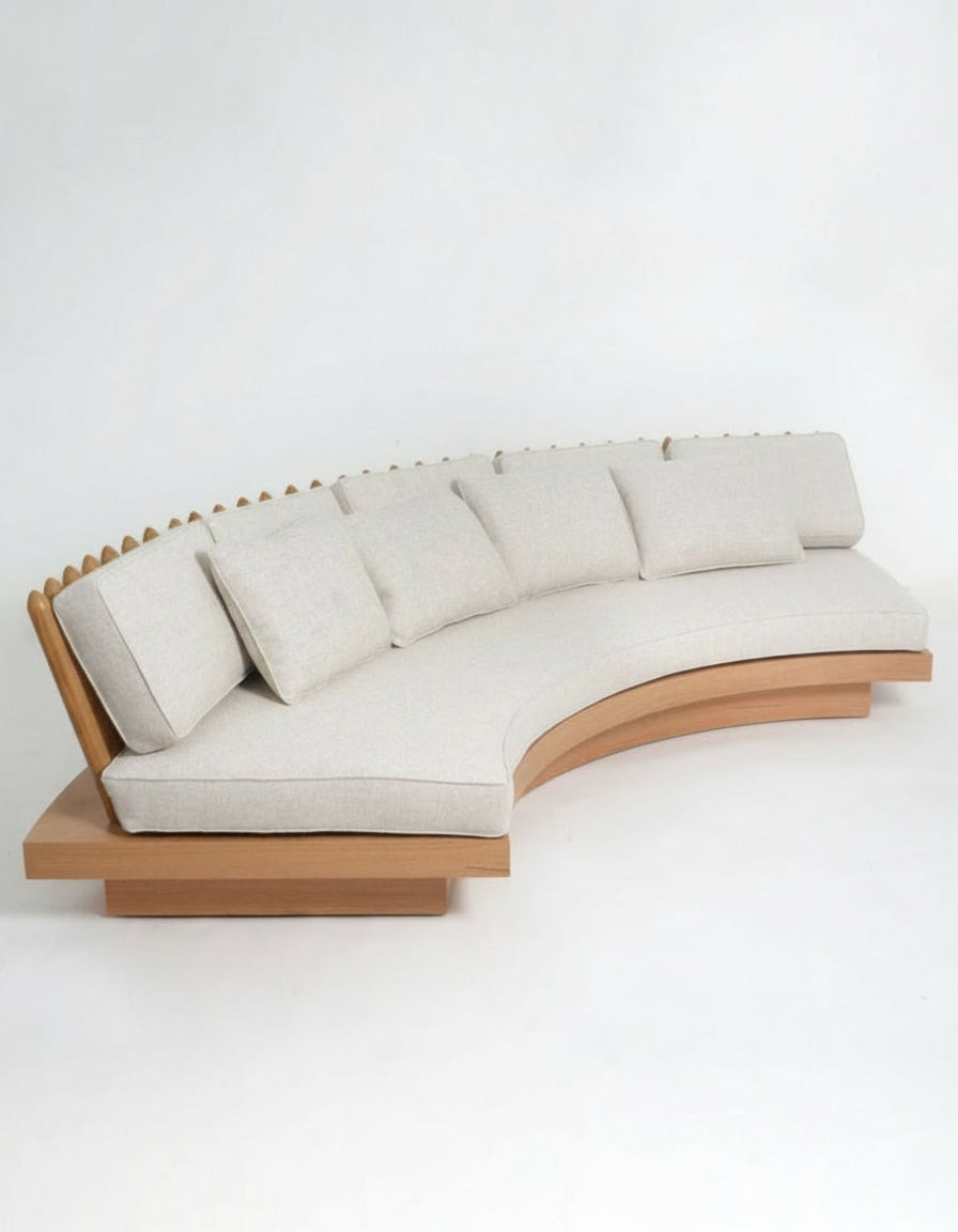 The San Romano Round Oak Sofa by Barracuda Interiors features a modern curved design with light beige cushions, a wooden base and backrest, and multiple square pillows for comfort. Shown against a plain white background.