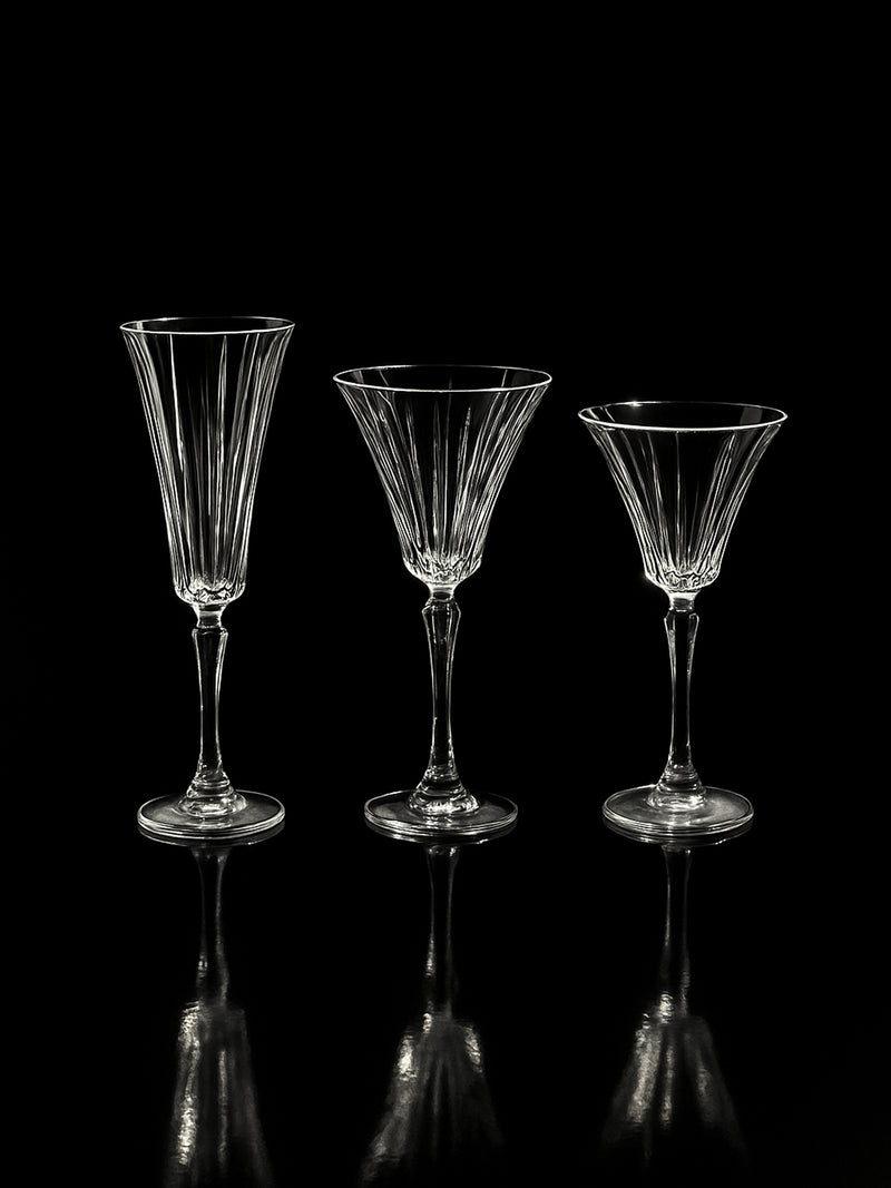 Three empty, elegant Dodo Vintage Italian champagne and wine crystal glasses and a carafe are arranged side by side against a black background, their hand-cut surfaces subtly reflecting on the glossy surface below.