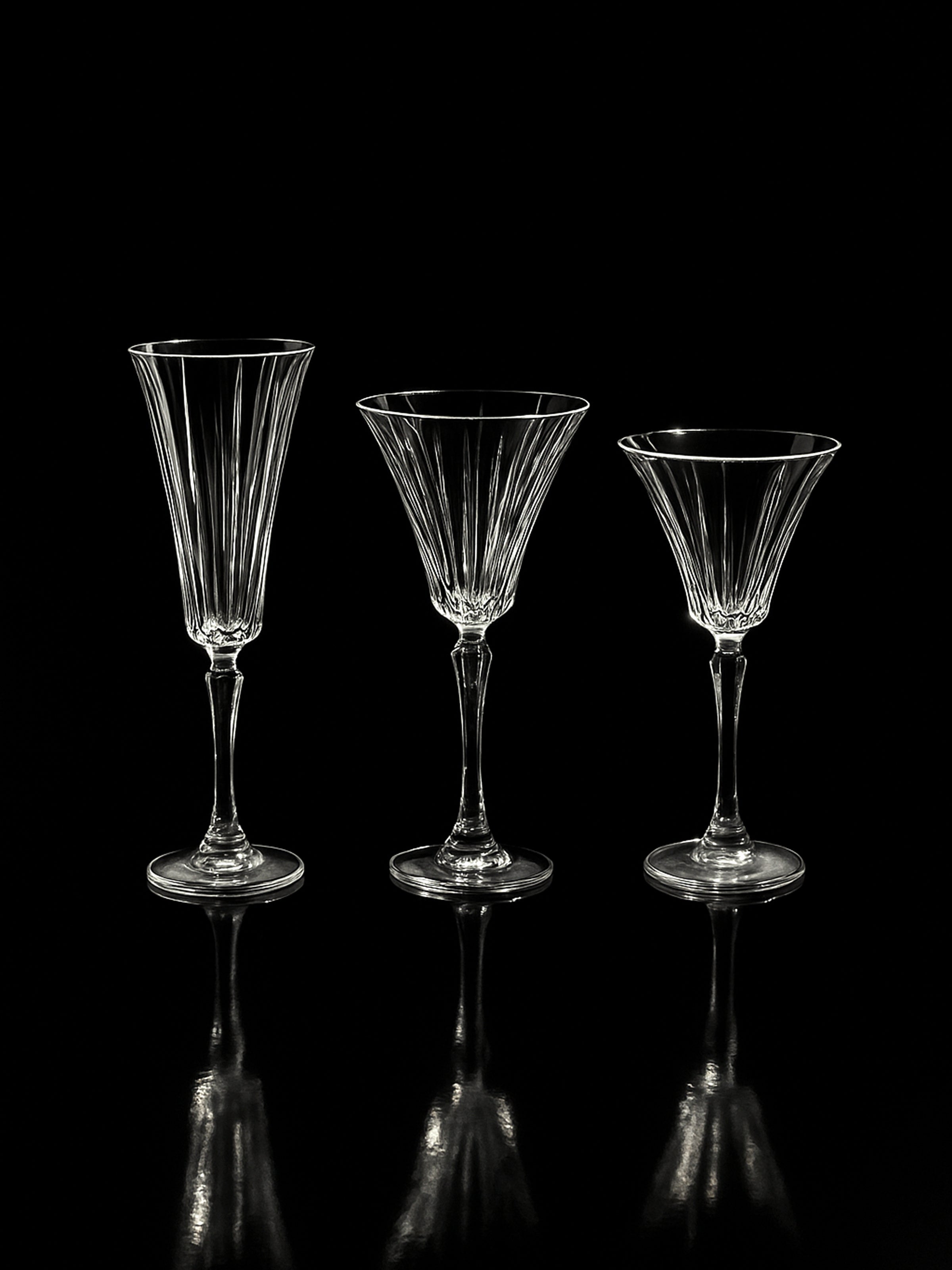 Three empty, elegant Dodo Vintage Italian champagne and wine crystal glasses and a carafe are arranged side by side against a black background, their hand-cut surfaces subtly reflecting on the glossy surface below.