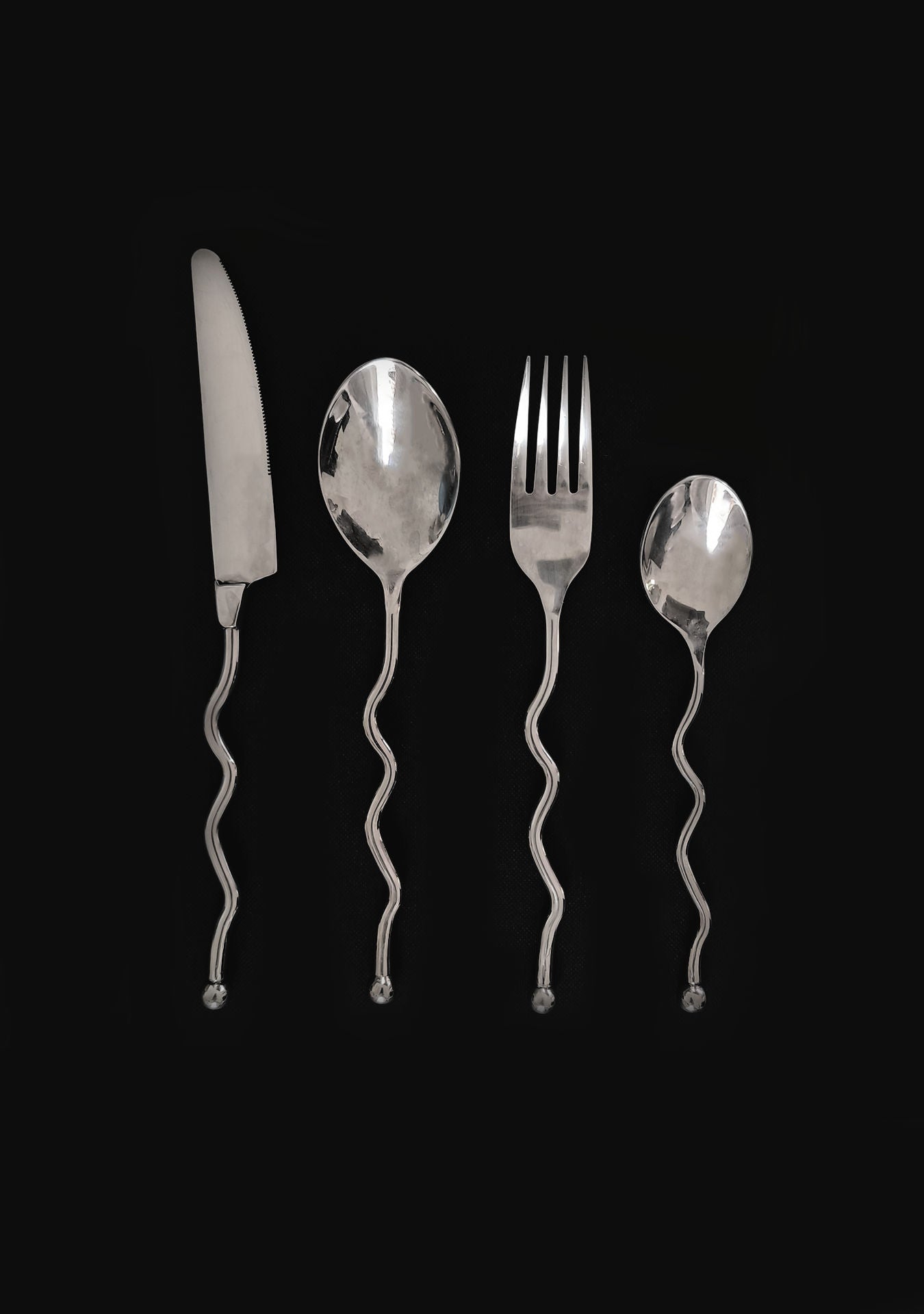 The Les Objoies Set of 4 Zig-zag Cutlery, featuring a knife, fork, spoon, and small spoon with wavy handles, is displayed in a row on a black background.