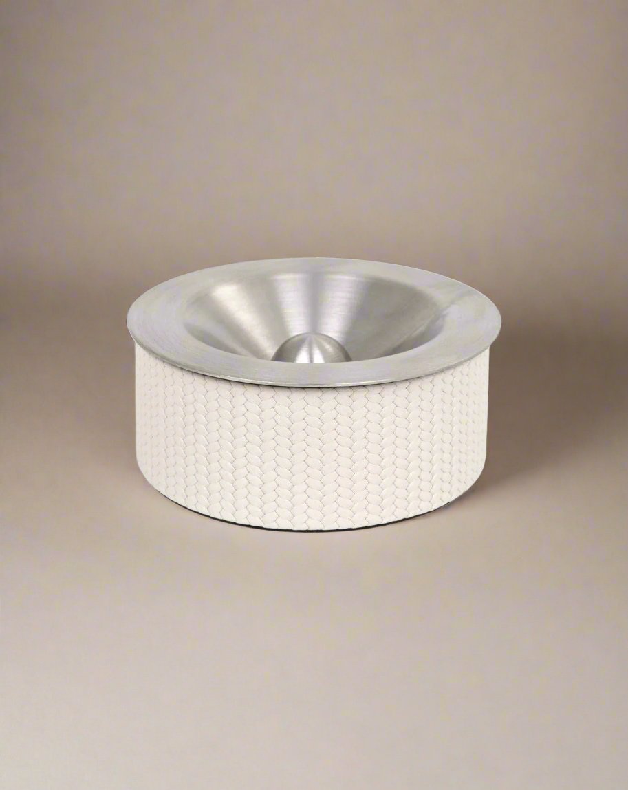 The Vulcano Ashtray by Pinetti is a contemporary art piece with a handcrafted, stainless steel, funnel-shaped top and a textured white base, set against a neutral background.