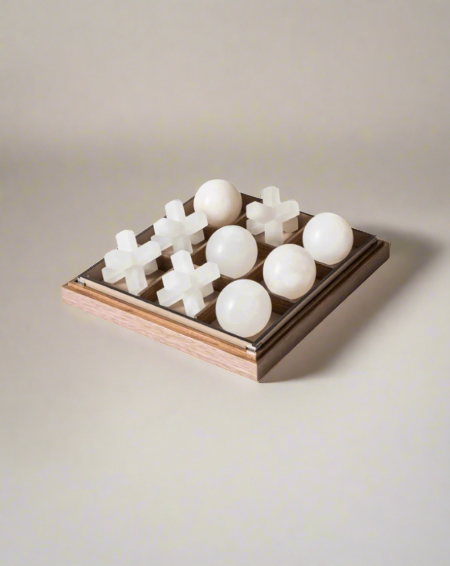 A Pinetti Tic Tac Toe game board with spherical white pieces and cross-shaped translucent pieces set on a luxurious Volterra alabaster board against a white background.