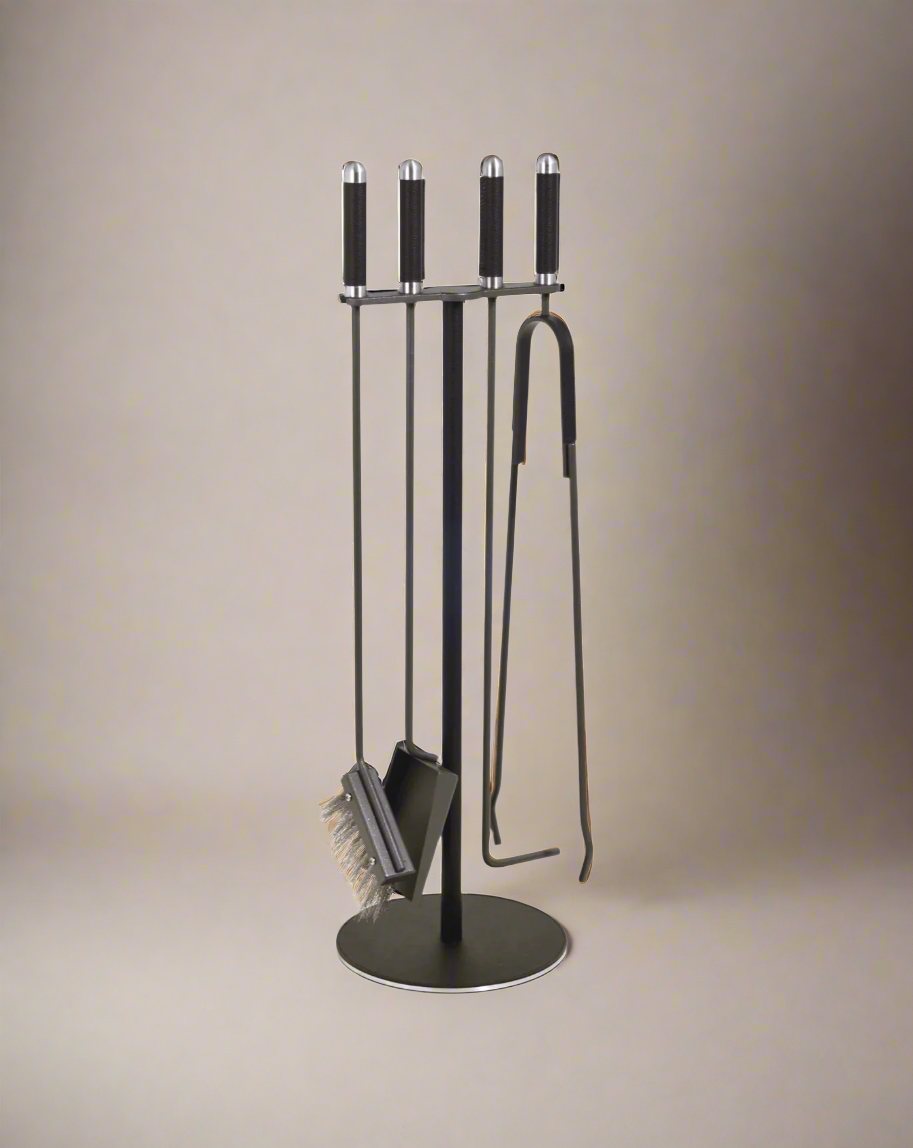 A Pinetti modern fireplace tool set in burnished steel, including a poker, brush, shovel, and tongs, neatly organized on a circular stand.