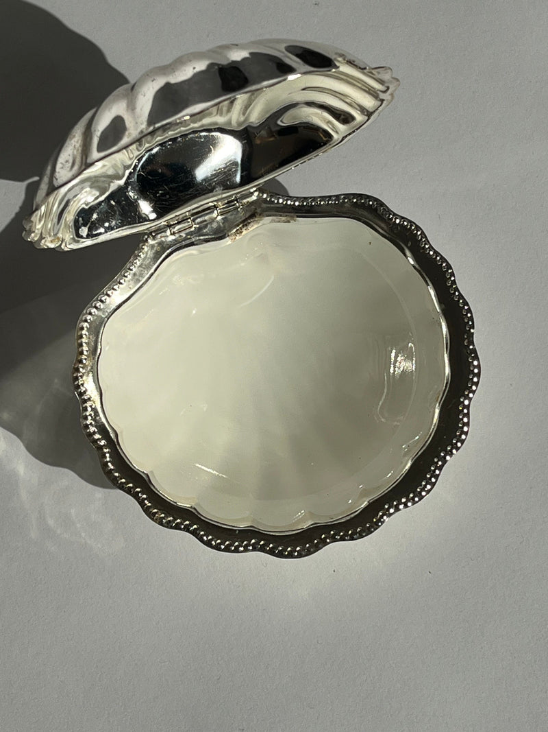The Shell Shaped Butter Dish by septembre studios features a shiny silver finish and hinged lid, revealing a smooth, empty interior and casting a soft shadow on a white surface.