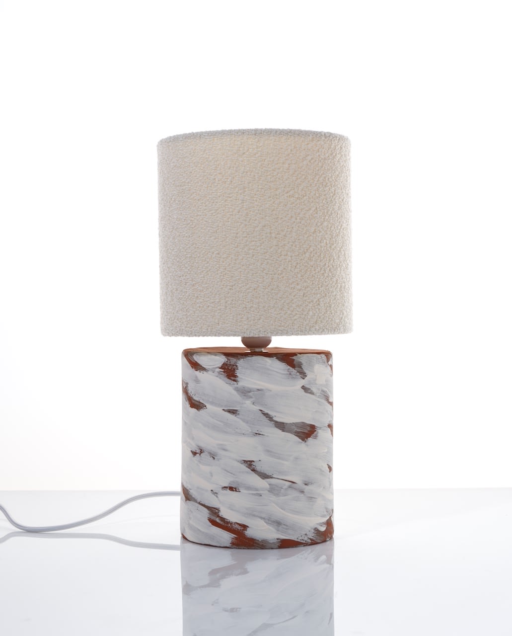 The Katia Langeard White Wind Table Lamp features a ceramic cylindrical base with white and brown brushstrokes and a textured off-white shade, ideal as a bedside lamp, shown on a glossy surface against a white background.