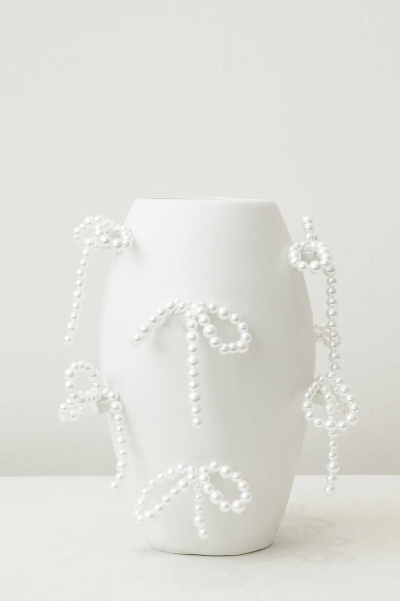 The "Notsobig" Power, Corruption, Lies Small Vase in matte white by Completedworks features faux pearl bows, showcasing distinctive decorative ceramics against a simple light backdrop.