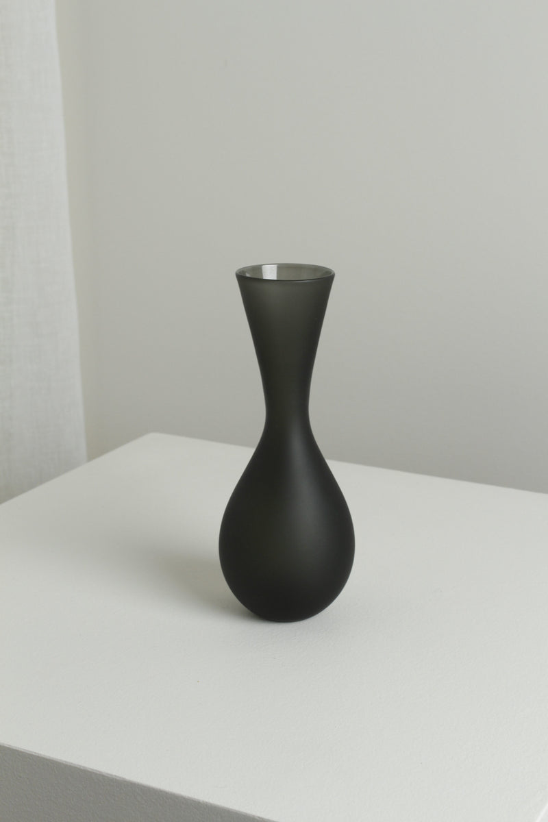 The Medium Vase in Smoke by Verre d'Onge, featuring an hourglass shape and matte black finish, is displayed on a white surface with a light gray background and softly lit white curtain to the left.