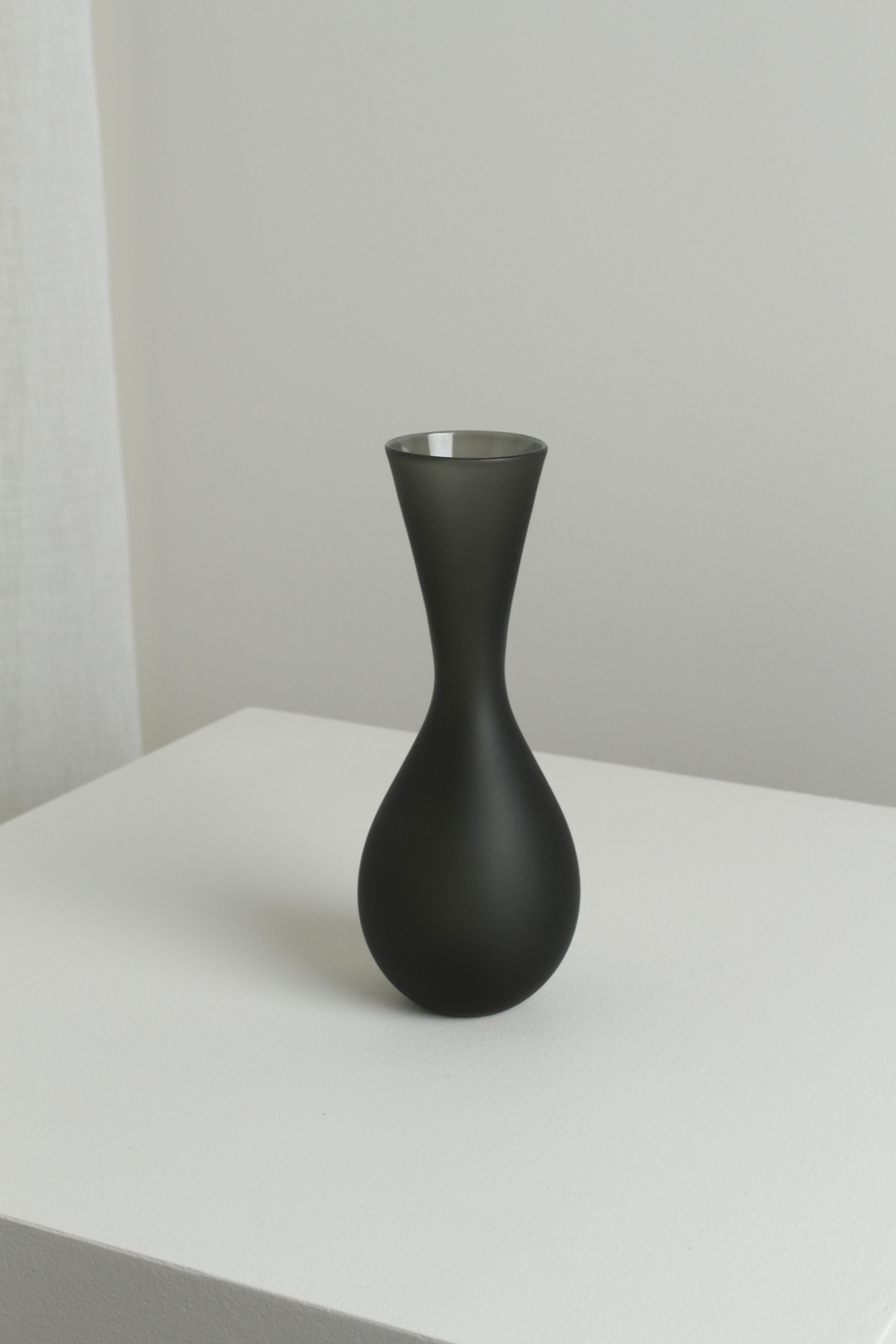 The Medium Vase in Smoke by Verre d'Onge, featuring an hourglass shape and matte black finish, is displayed on a white surface with a light gray background and softly lit white curtain to the left.