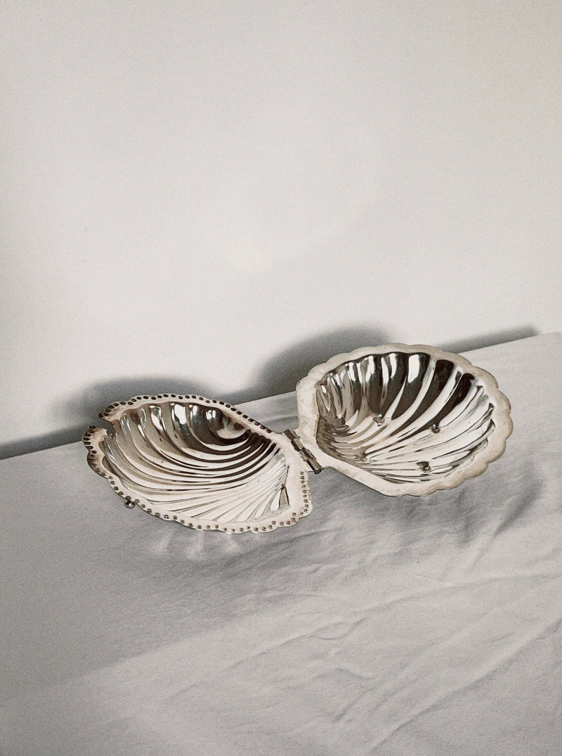 The Manushi Shell Shaped Butter Dish, with ridged reflective interiors and delicate curved edges, sits open on a white surface against a plain off-white background.