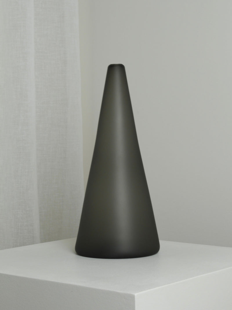 The XLarge Vase in Smoke by Verre d'Onge, featuring a cone shape and matte black finish, sits on a white pedestal in a minimalist room with white walls and a sheer curtain.
