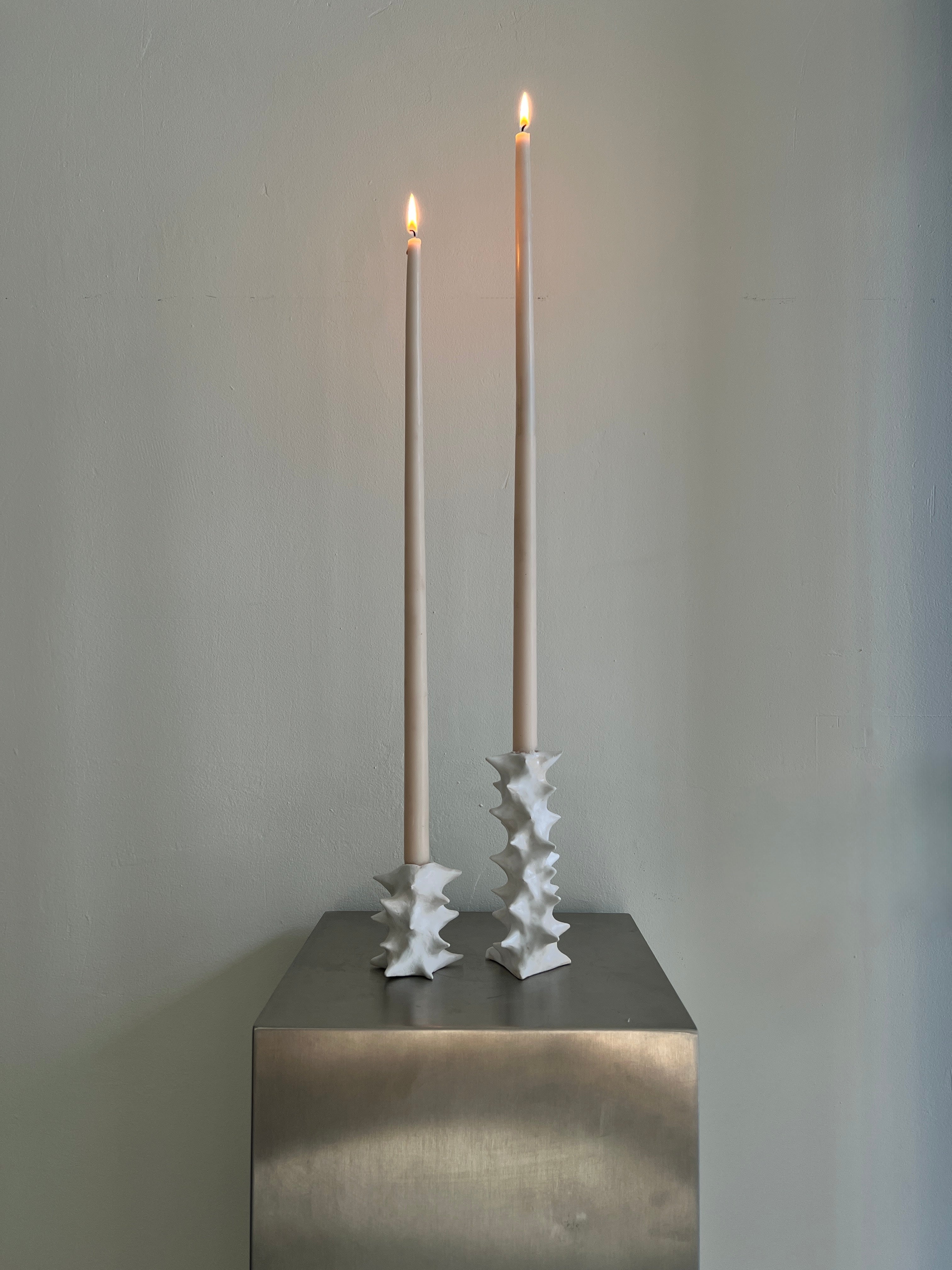 Two tall, lit candles sit in 2222Studio Spiky ceramic candle holders, crafted from matte white stoneware, on a metallic rectangular pedestal against a plain light gray wall.